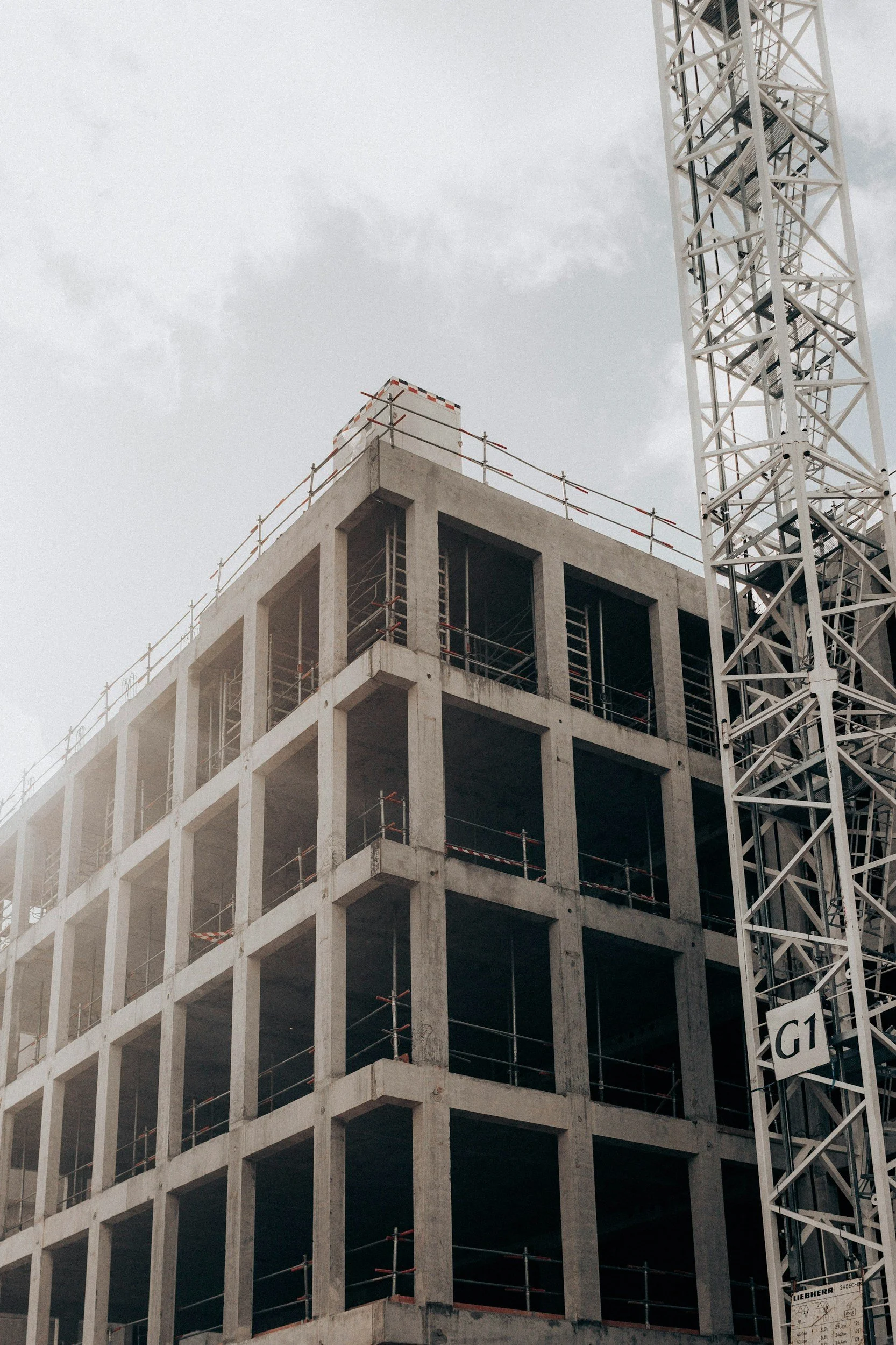 Construction site with a high-rise building under construction, featuring a skeletal concrete structure with open floors and safety railings. A large white construction crane is positioned to the right of the building.