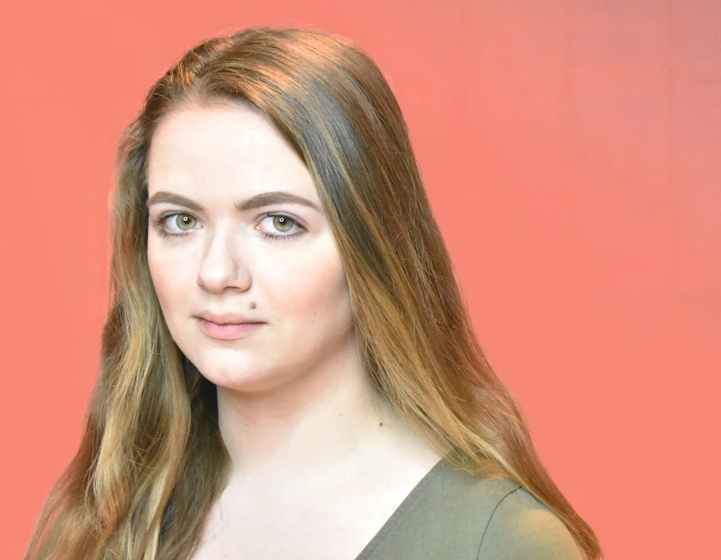A young woman with long, reddish-brown hair and hazel eyes posing against a pinkish-orange background.
