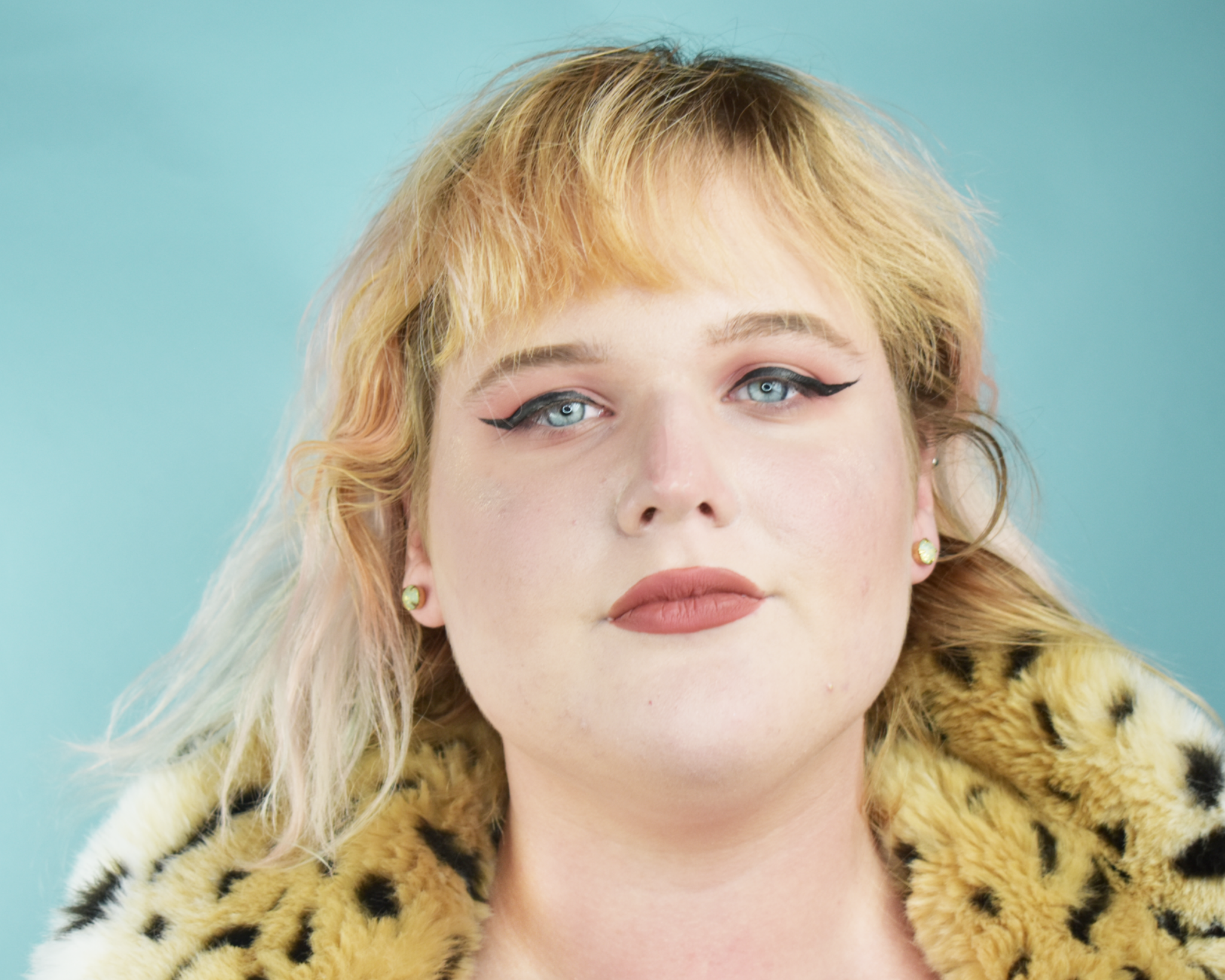 Close-up of a person with blonde hair, wearing bold black eyeliner, gold earrings, and a leopard-print garment against a light blue background.
