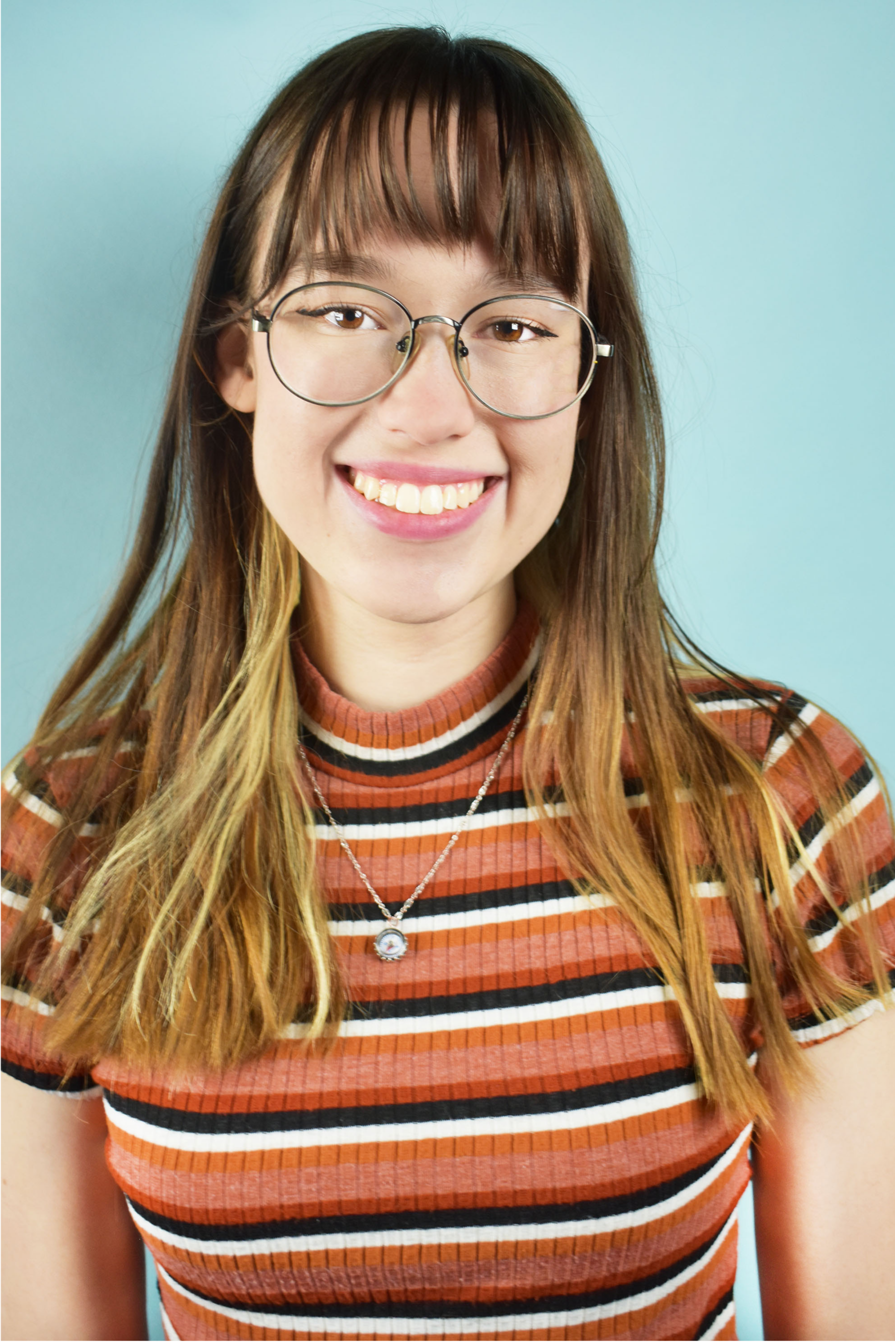 Close-up of a smiling young woman with glasses, long wavy hair, wearing a striped shirt and a necklace, against a light blue background.