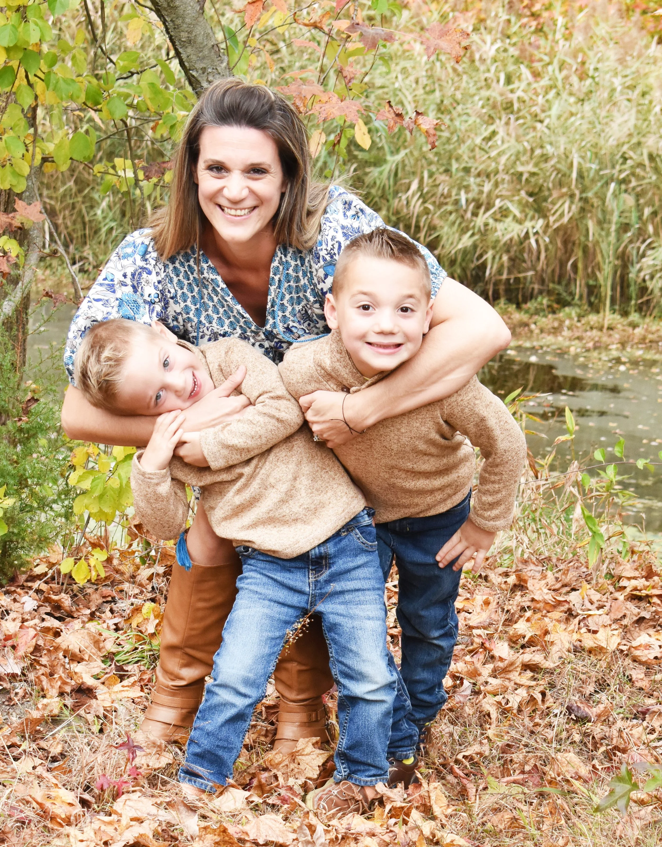 A woman with brown hair smiling and hugging two young boys outdoors among fall foliage near a pond.