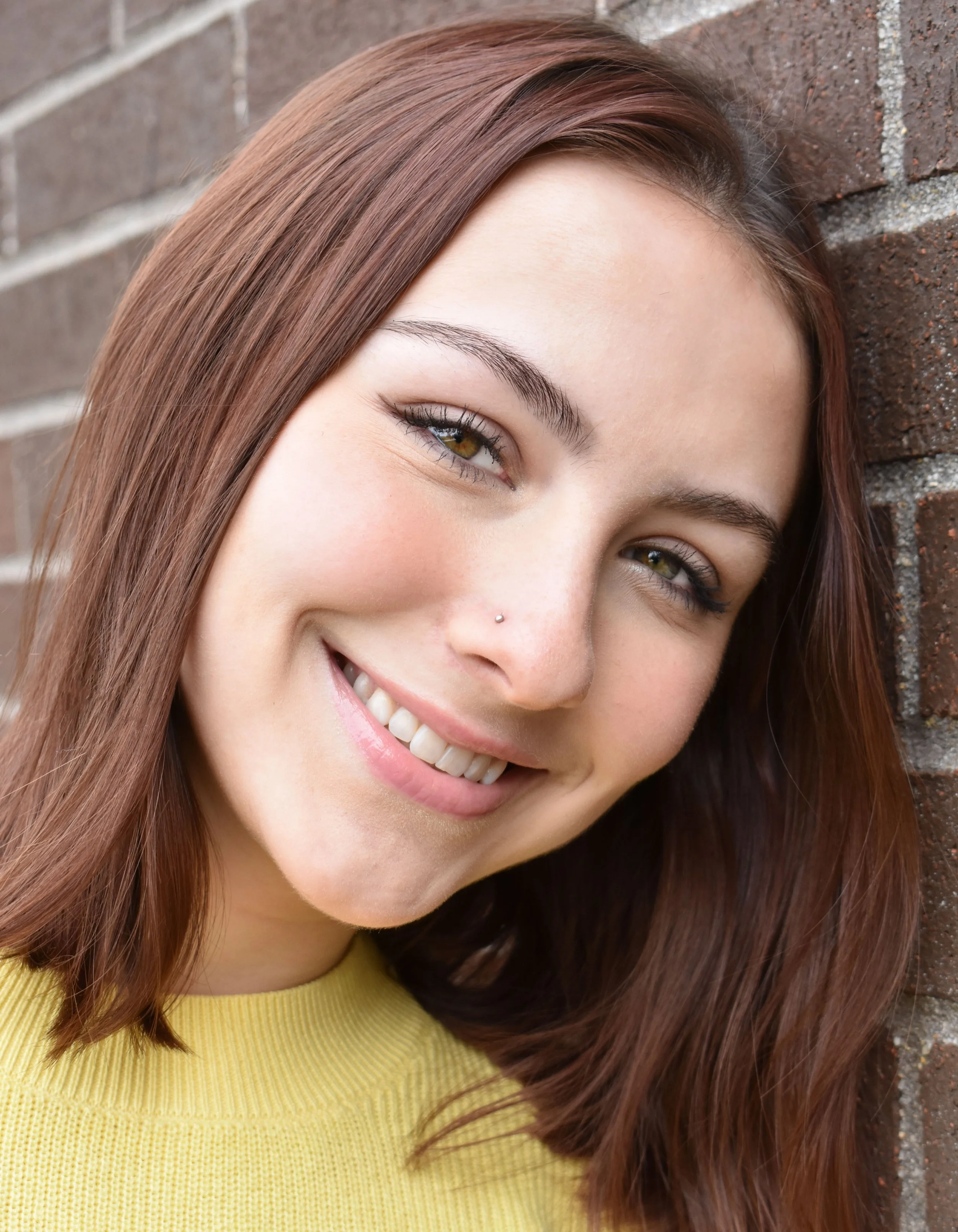 A smiling woman with long brown hair and hazel eyes, wearing a yellow sweater, standing against a brick wall.