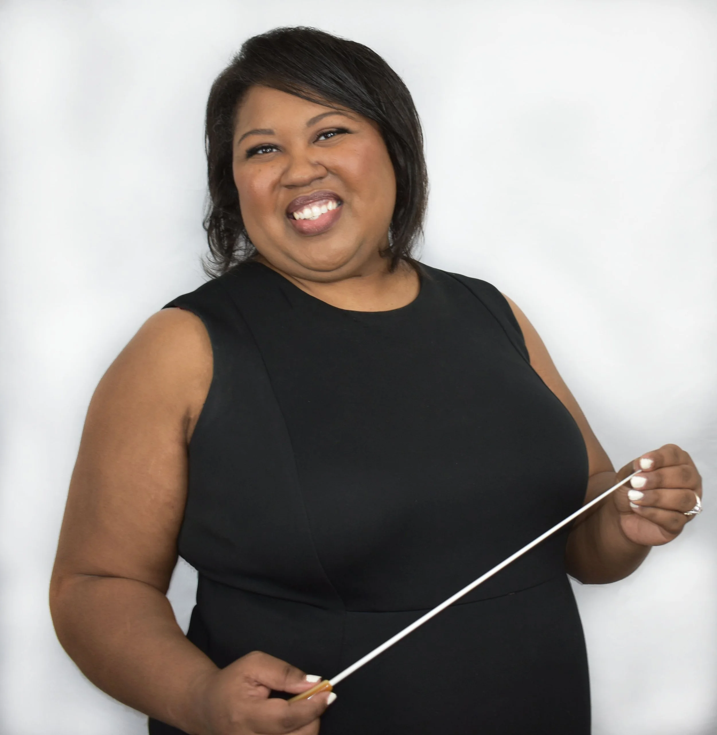A woman with dark hair in a bob haircut wearing a black sleeveless top, smiling and holding a disposable wooden stick, standing against a plain white wall.