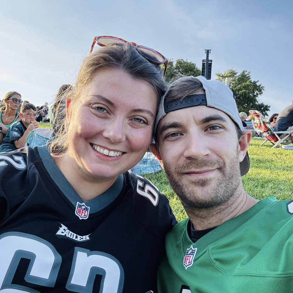 A smiling couple taking a selfie outdoors at a sports event, wearing NFL football jerseys, with people sitting on grass and tents in the background.