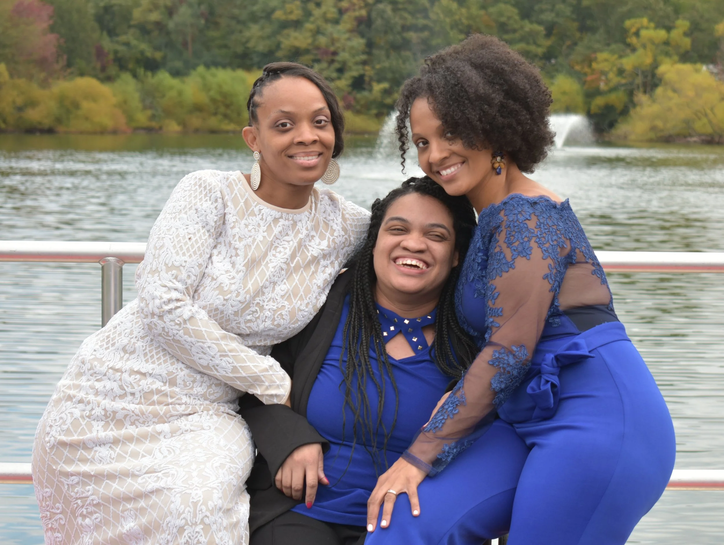 Three women smiling together outdoors near a lake with trees in autumn colors in the background.