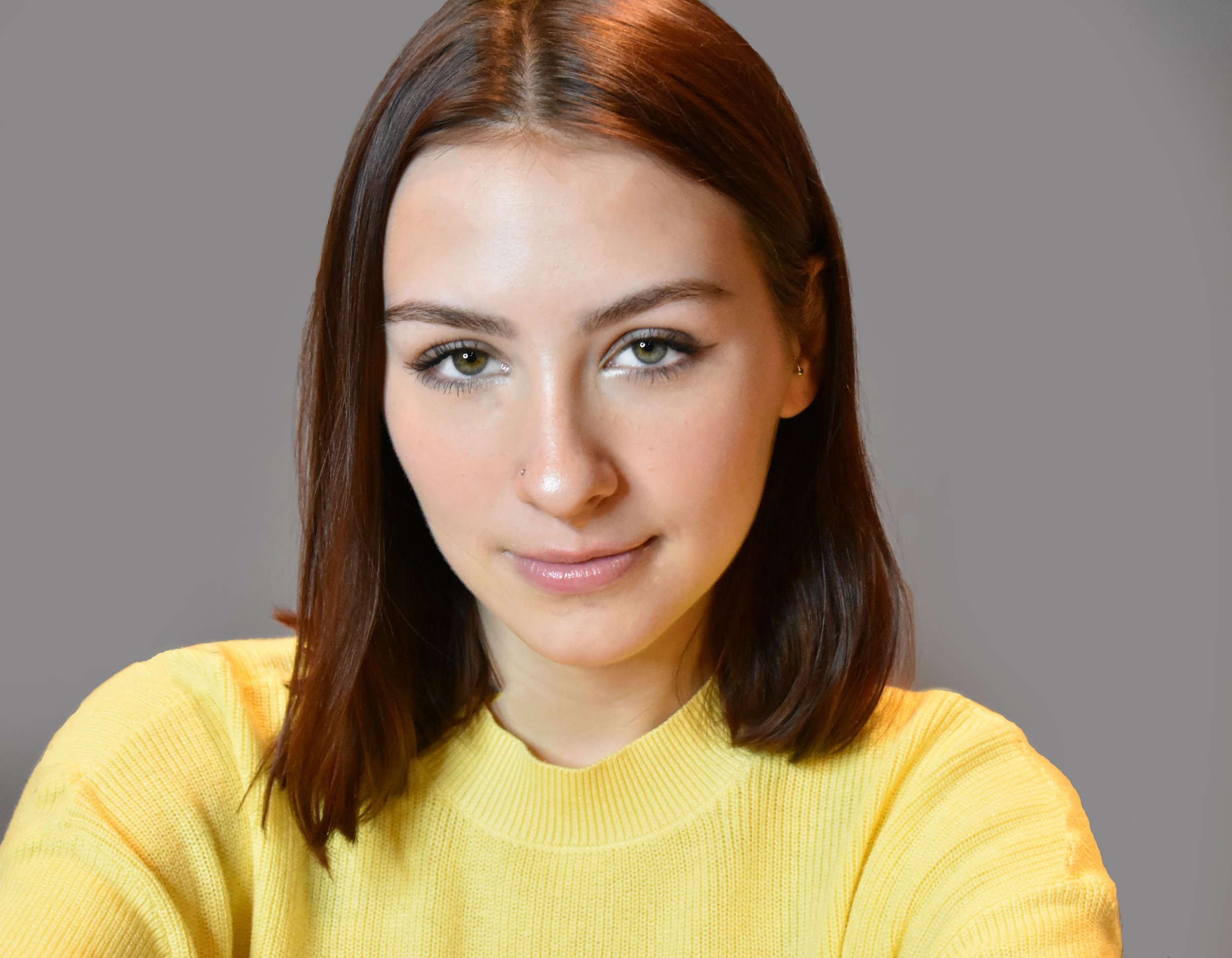 Close-up of a young woman with red hair, hazel eyes, and a nose piercing, wearing a yellow sweater, smiling softly against a gray background.
