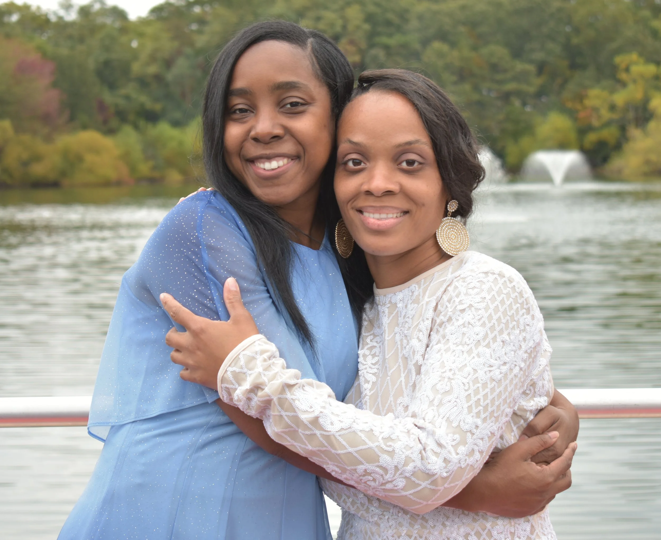 Two women hugging each other outdoors near a body of water with fountains, surrounded by trees with autumn foliage.