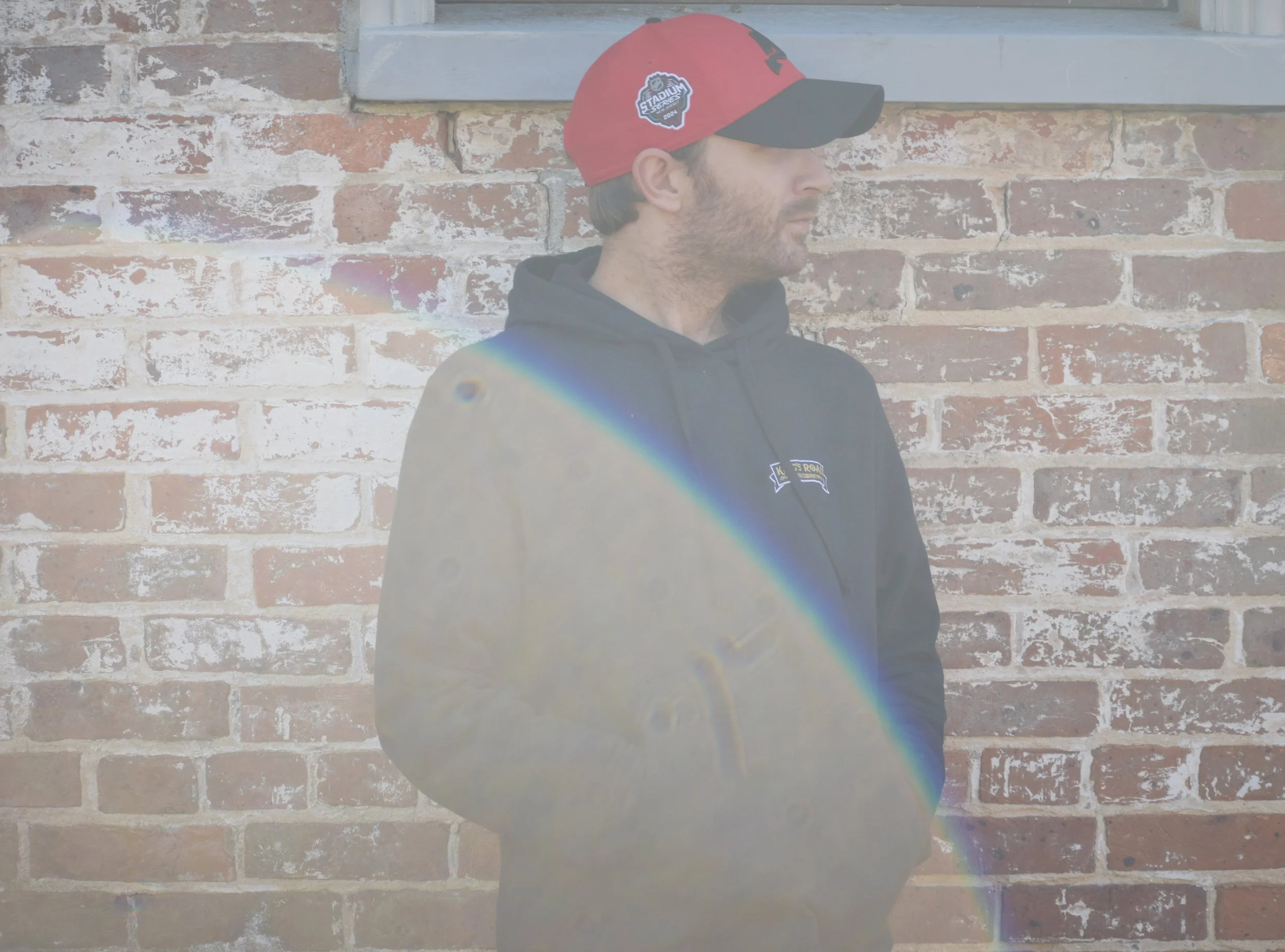 A man wearing a red and black baseball cap and a black hoodie, standing against a brick wall, with a rainbow-colored light streak crossing his chest.