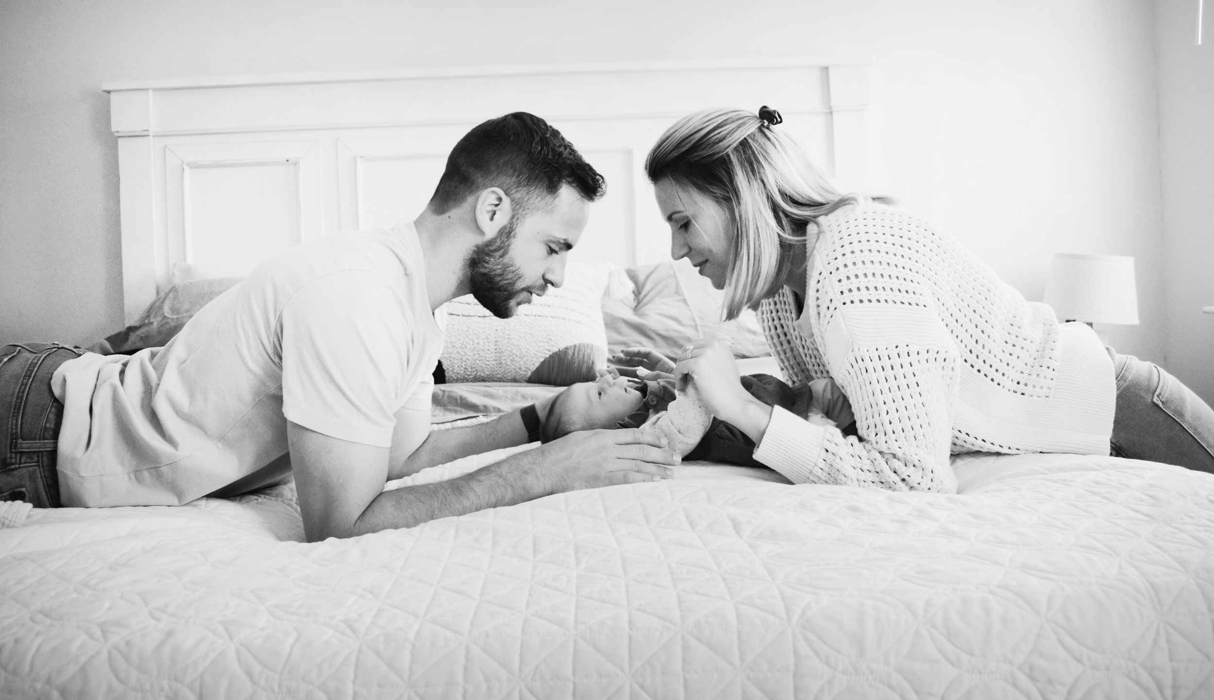 Black and white photo of a young family on a bed, with a father and mother leaning close to their baby, smiling and holding the baby's hands.