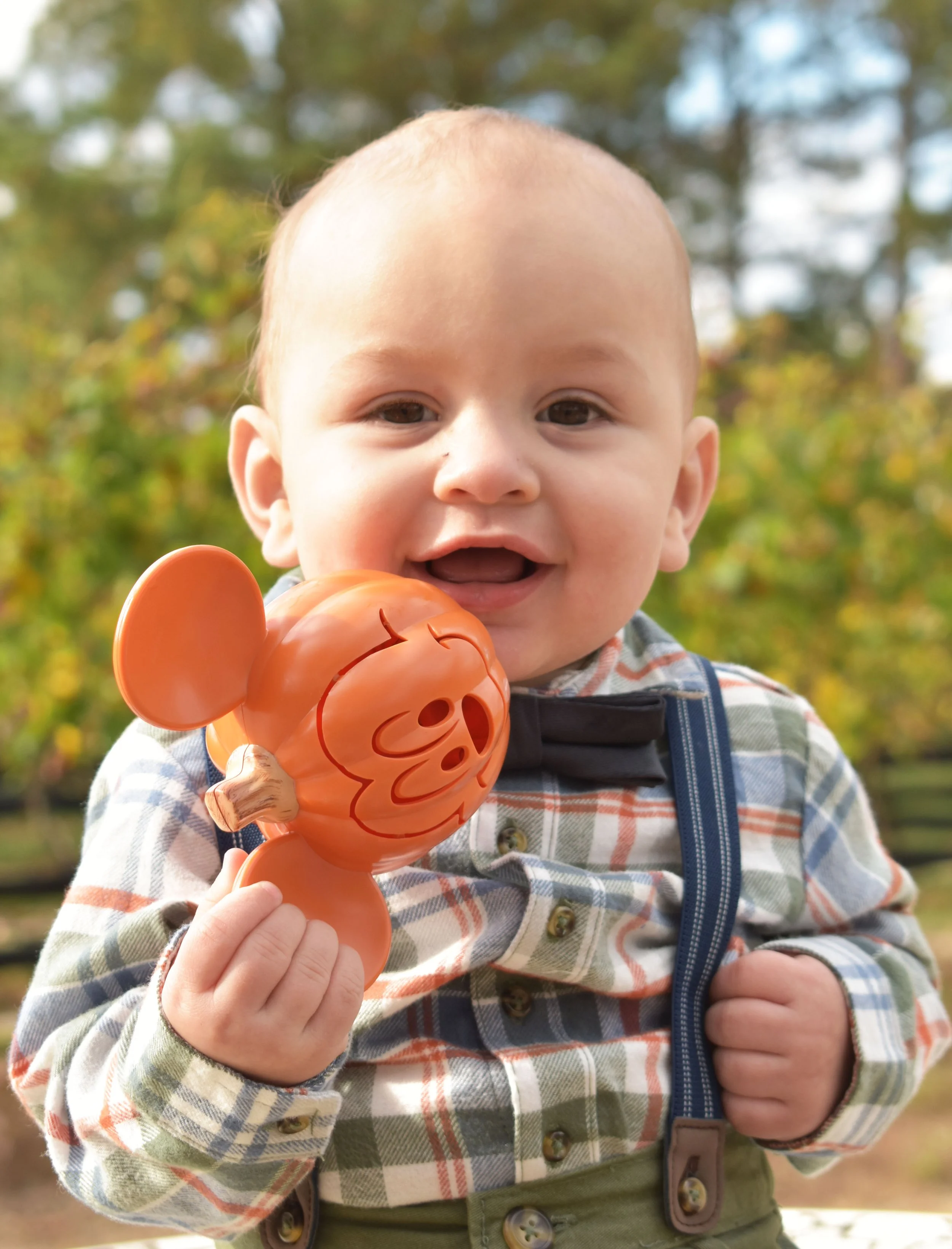 Close-up of a smiling baby holding a pumpkin-shaped toy outdoors with trees in the background.