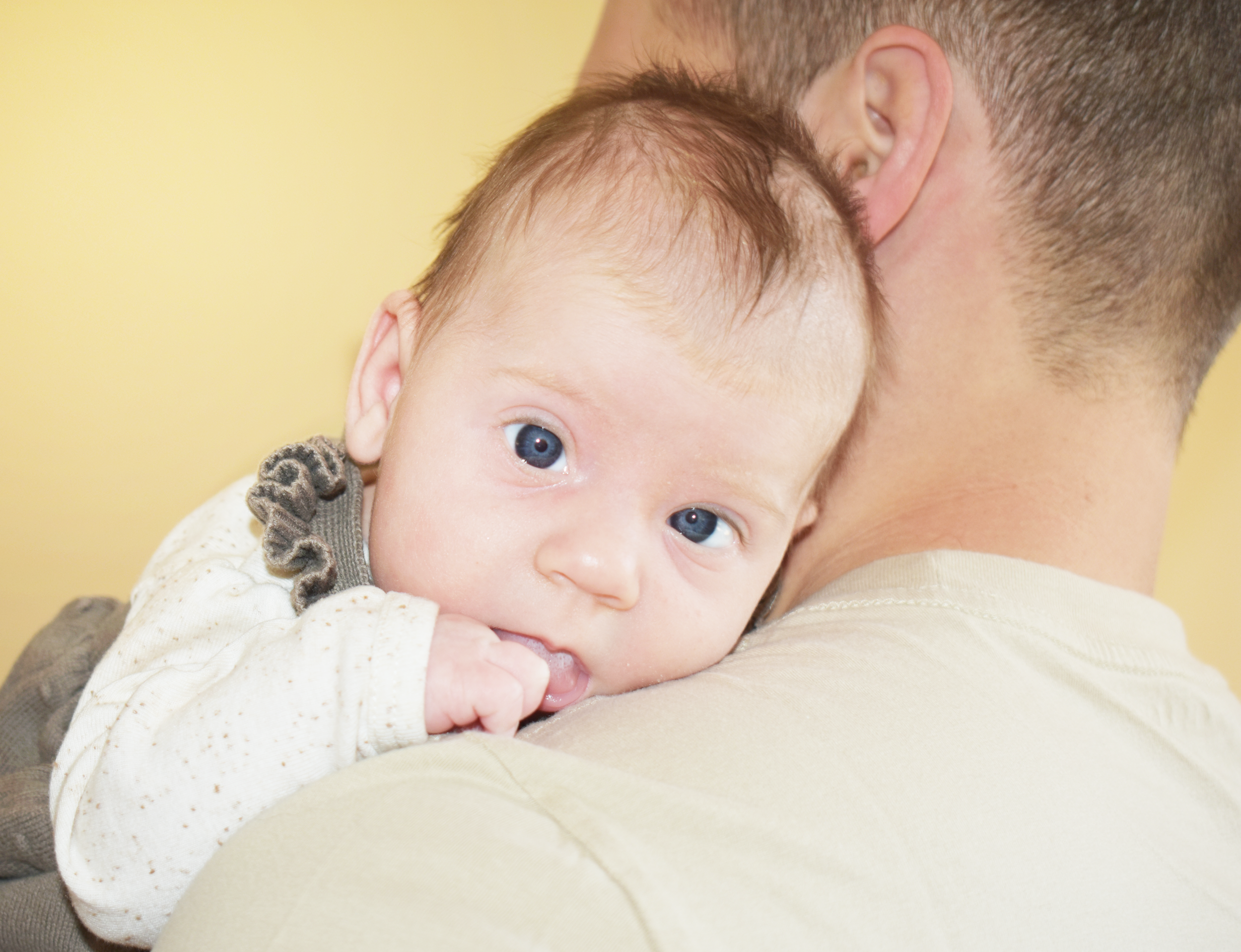 A close-up of a young child with blue eyes, lying on an adult's shoulder, with an adult's neck and ear visible, set against a plain yellow background.