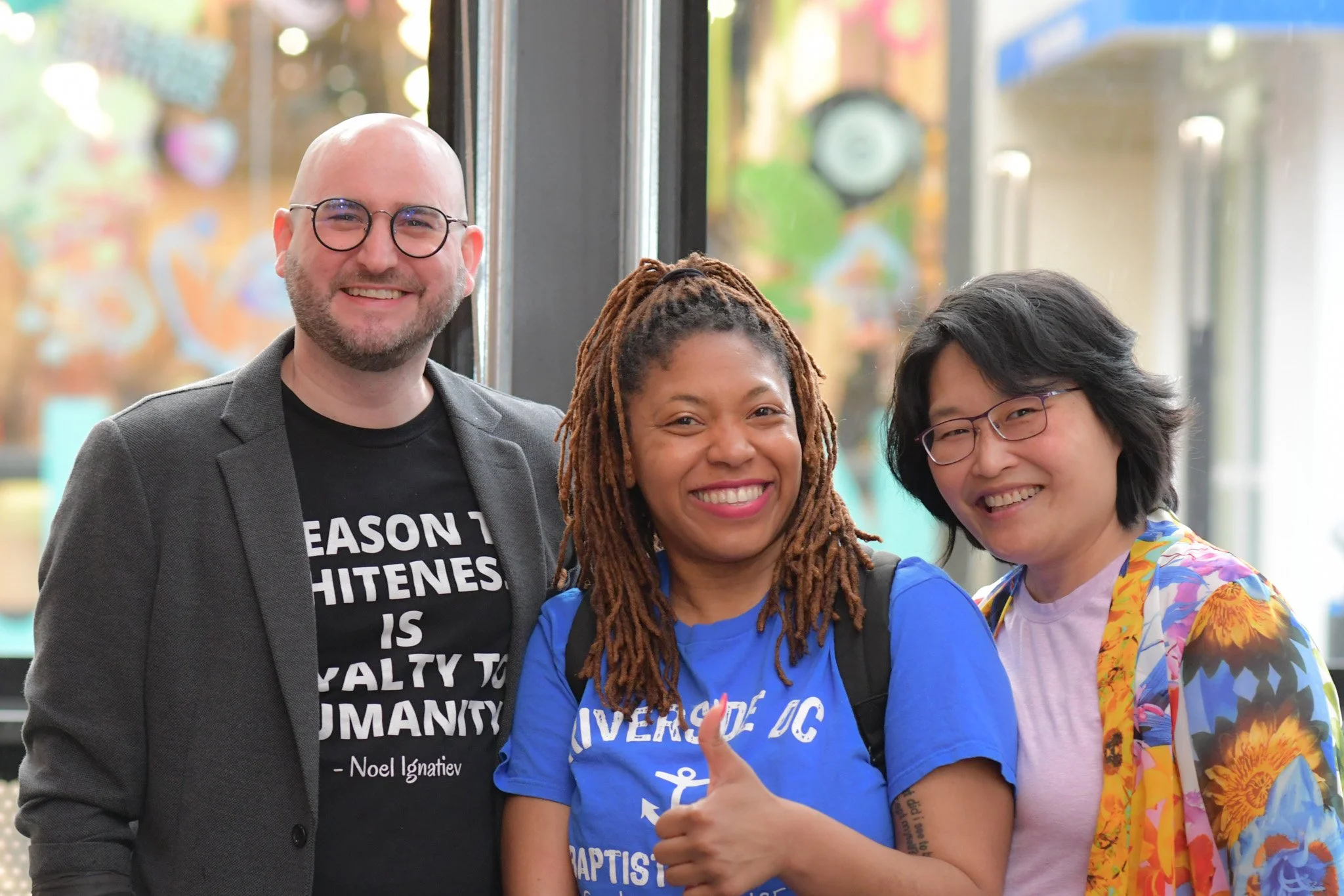A group of three diverse smiling people standing close together inside in front of large windows with blurred colorful outdoor scenery. The person on the left is a bald man wearing glasses, a dark blazer, and a black T-shirt. The woman in the middle has long, braided hair, a bright smile, and is wearing a blue T-shirt with white text. The person on the right has short, dark hair, glasses, and is wearing a colorful, patterned jacket with a pink T-shirt underneath.