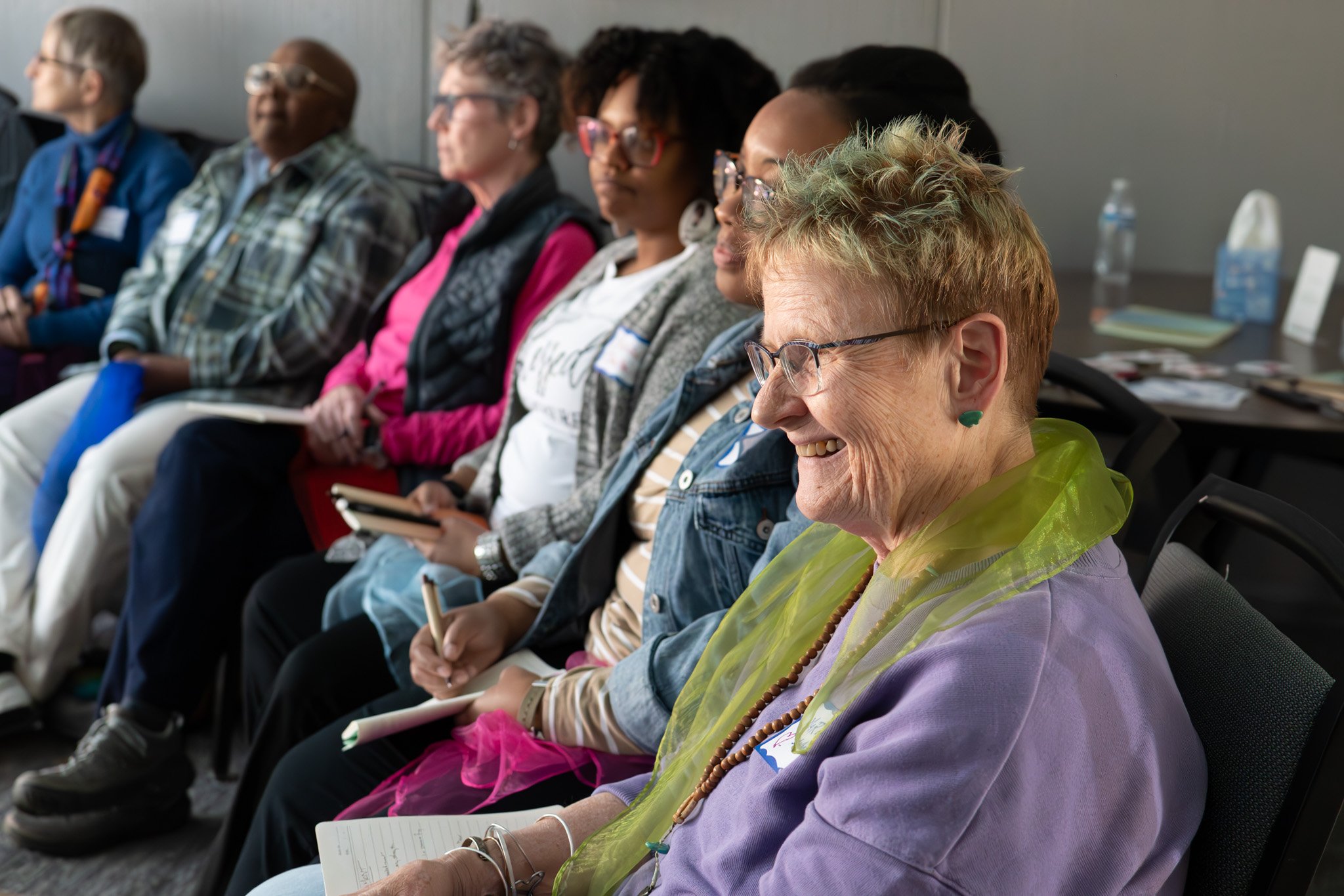 Group of diverse women seated in a conference room, listening and taking notes at a meeting or seminar.