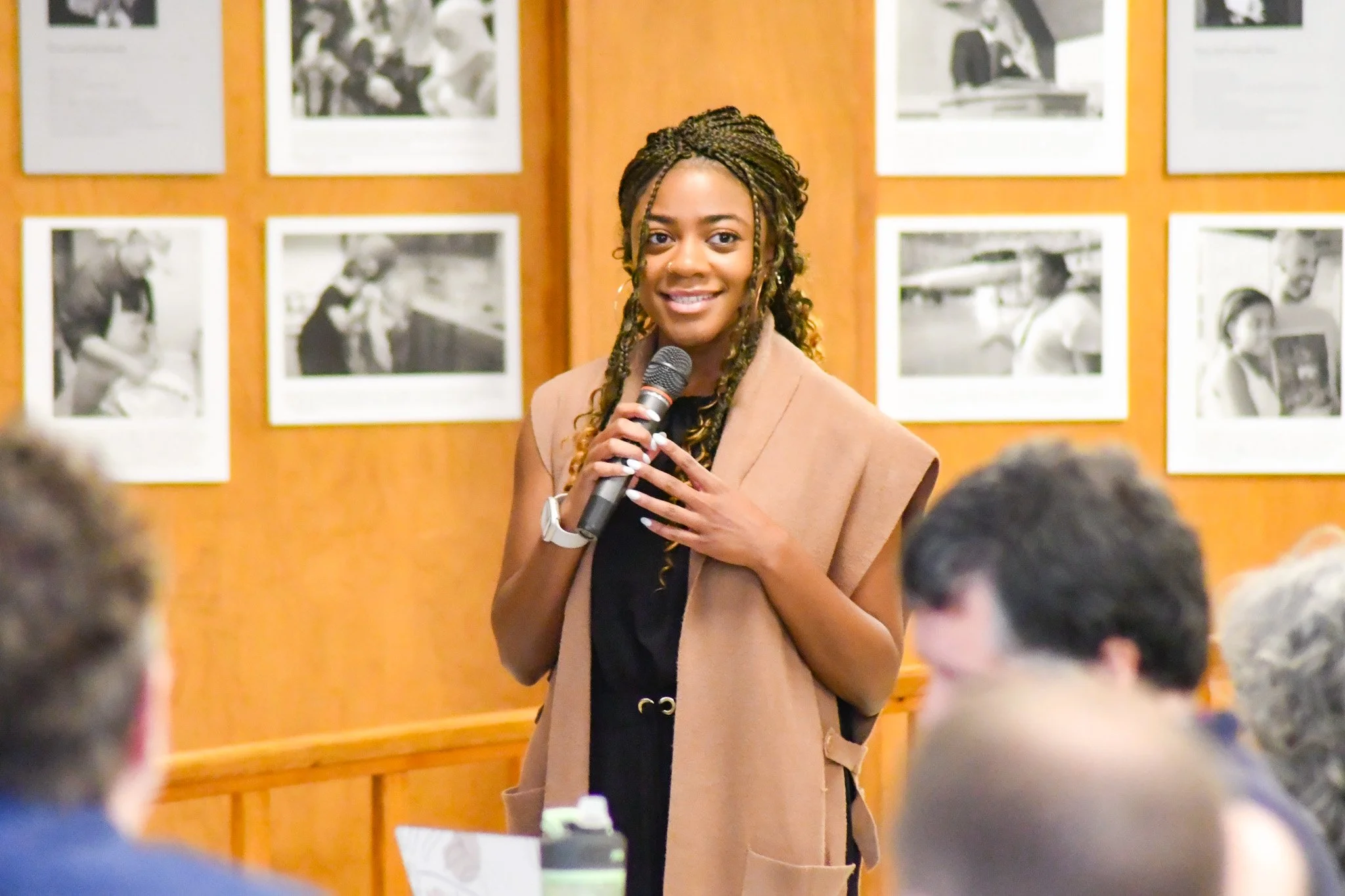 A young woman with braids smiling and holding a microphone speaks to an audience at an indoor event, with photographs displayed on the wall behind her.