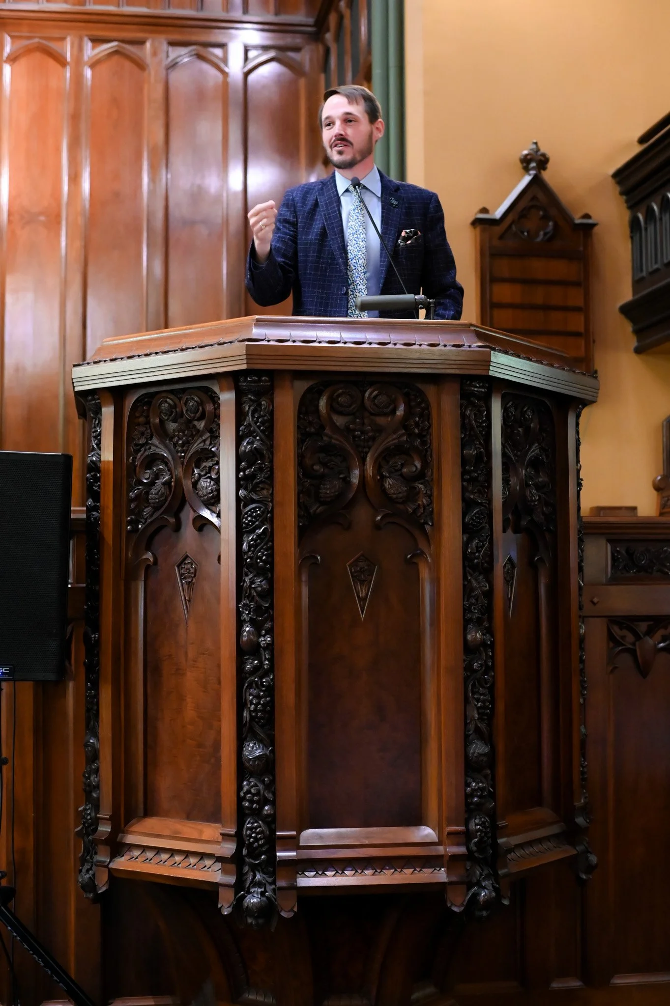 A man in a suit giving a speech at a wooden pulpit with intricate carvings, inside a room with wooden paneling and ornate woodwork.