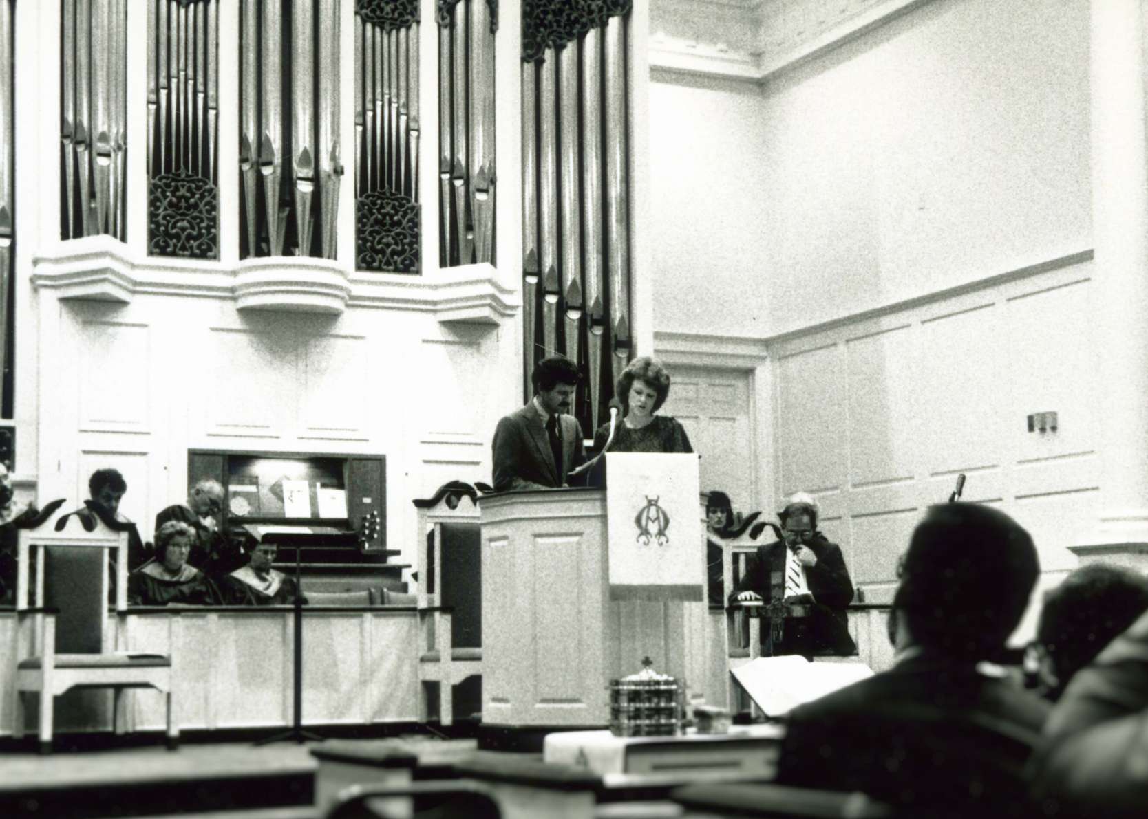 A black and white photo of a formal gathering or ceremony in an ornate hall with a large pipe organ in the background. Two women are speaking at a podium, with several seated individuals, including a man in a suit and tie, listening attentively.