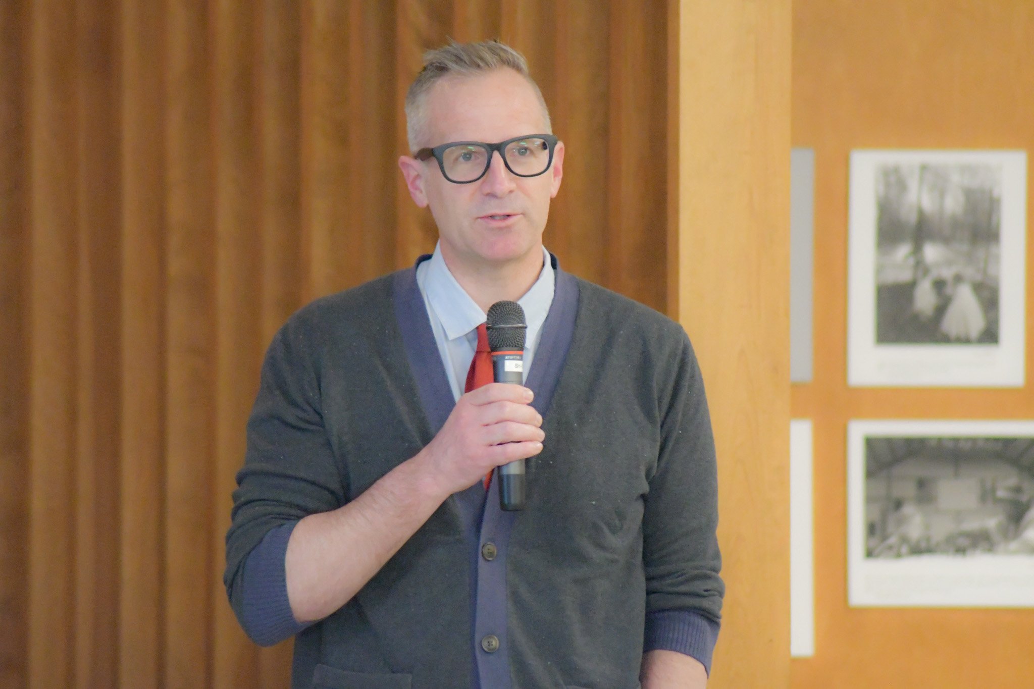 A man with glasses, short hair, and light skin holding a microphone, standing indoors with a wooden wall and framed pictures in the background.