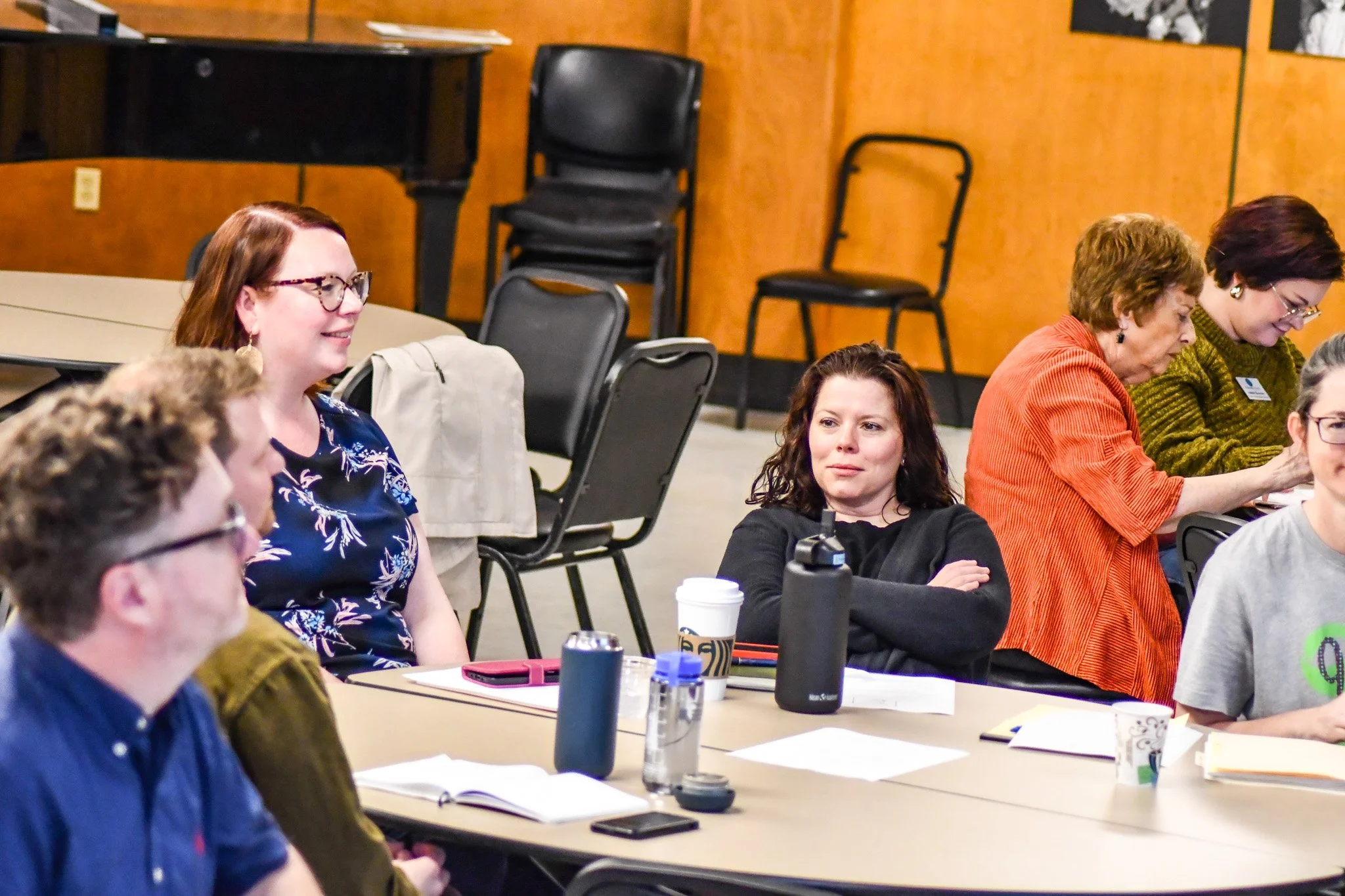 Group of women sitting at a long table in what appears to be a meeting or workshop. They are engaged, with notebooks, coffee cups, and water bottles on the table.