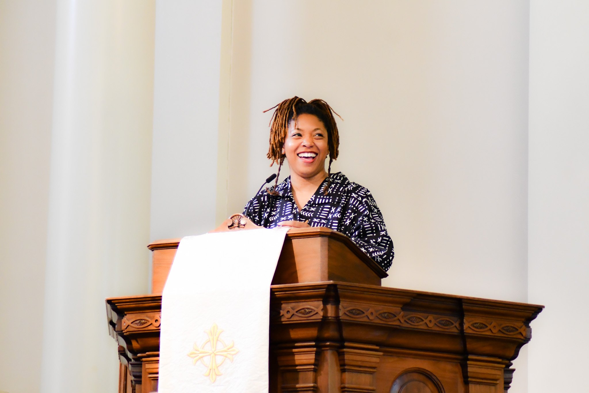A woman with dreadlocks and a black and white patterned shirt stands at a wooden podium inside a church or chapel, smiling.