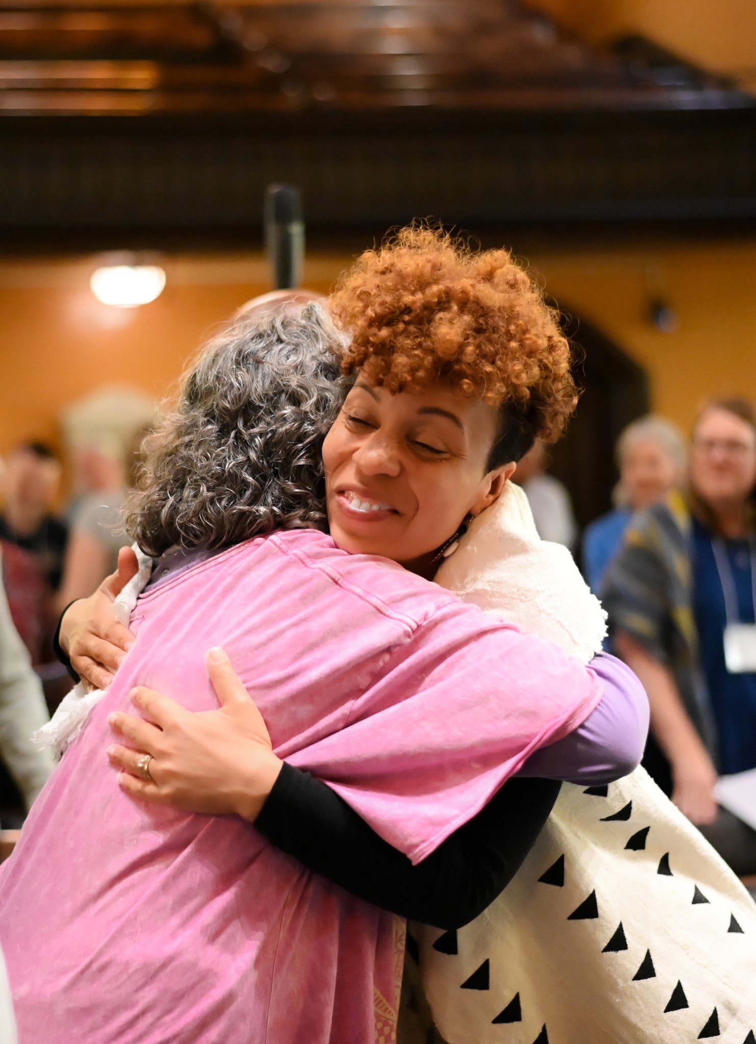 Two women hugging and smiling, one with curly ginger hair and the other with curly gray hair, in a warm indoor setting with blurred people in the background.