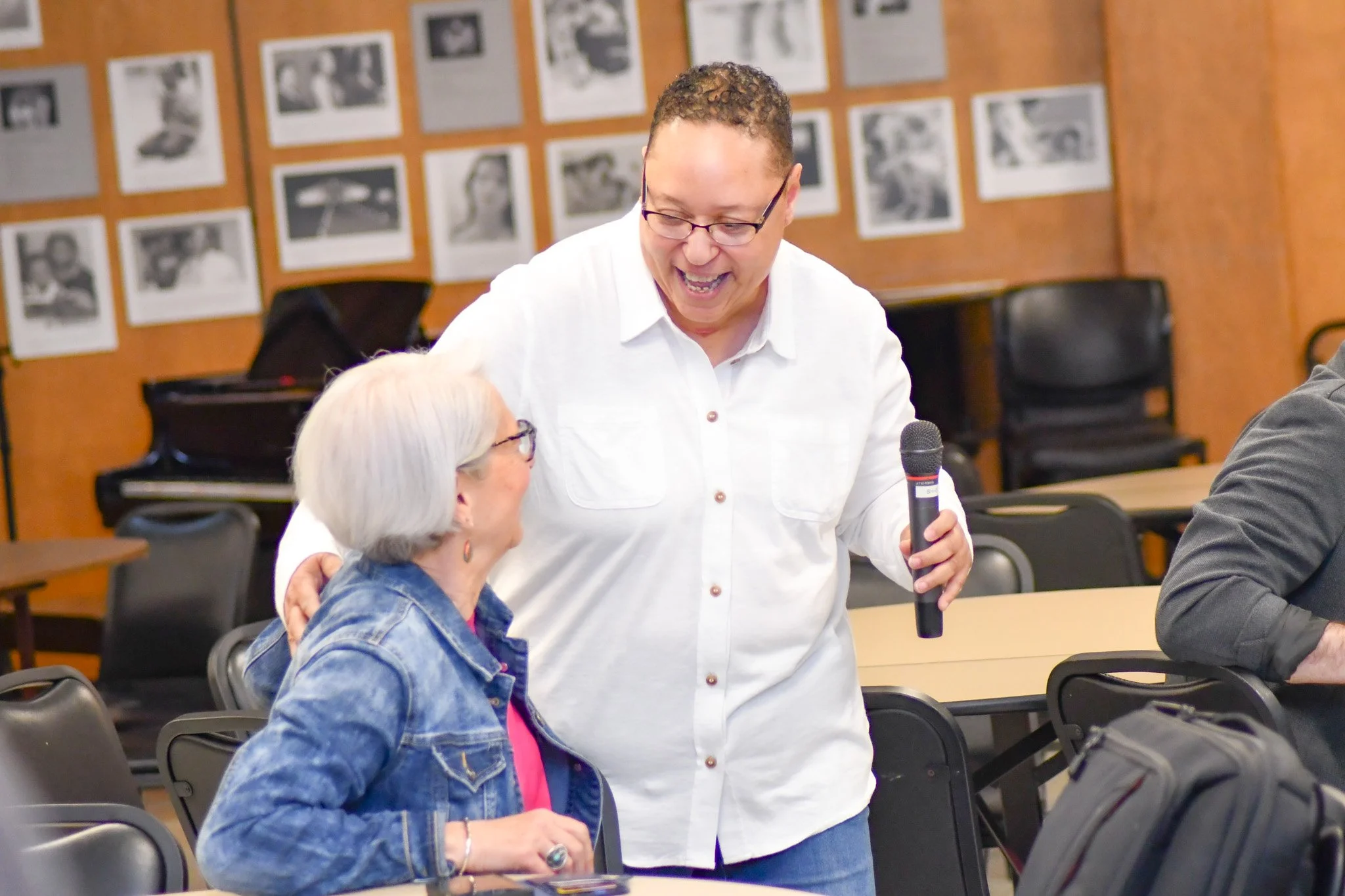 A man with curly hair and glasses, dressed in a white shirt, holding a microphone, smiling and engaging with an elderly woman with white hair, glasses, and a denim jacket, in a room with photographs on the wall.