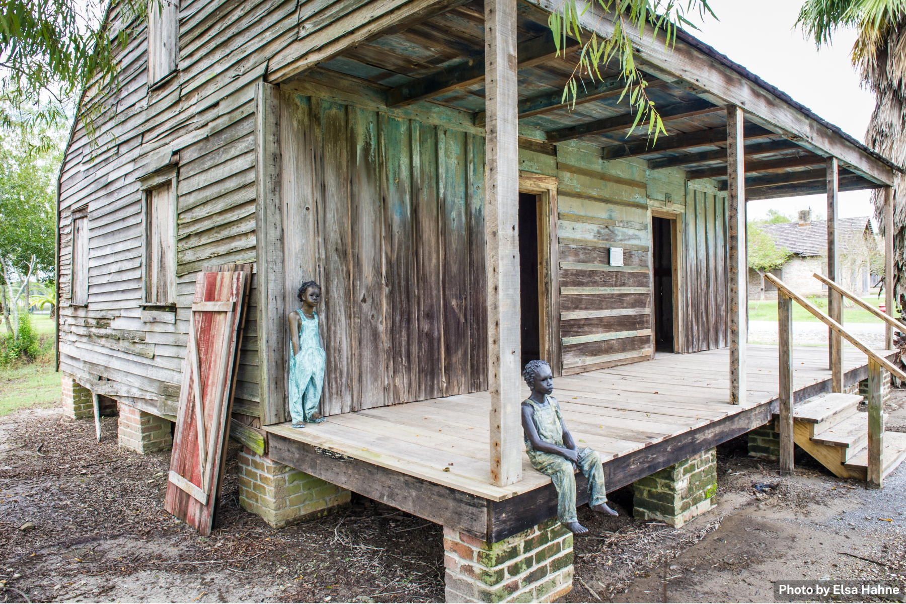 An old wooden house with a porch, featuring two seated metal statues of children and a standing statue of a girl in a dress. A painted wood panel is leaning against the house. The porch is elevated on brick pillars, with a path leading up to it, and the house is surrounded by trees and greenery.