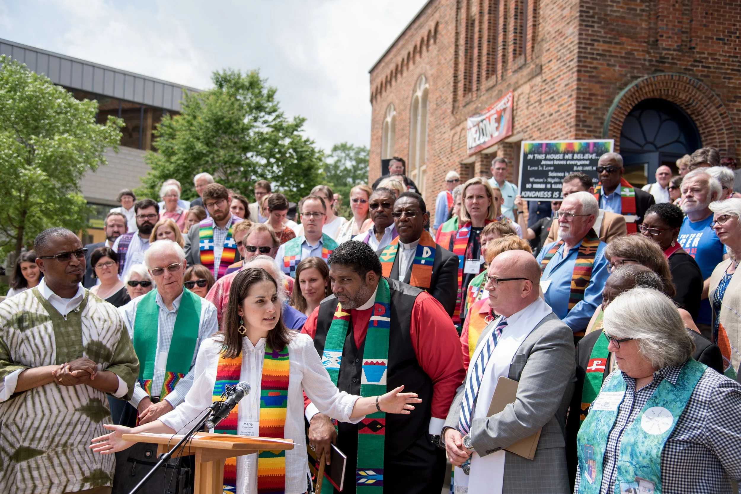 A diverse group of faith leaders gathered outside a brick building during a community event, some wearing rainbow-colored stoles and accessories, with a woman speaking at a podium.