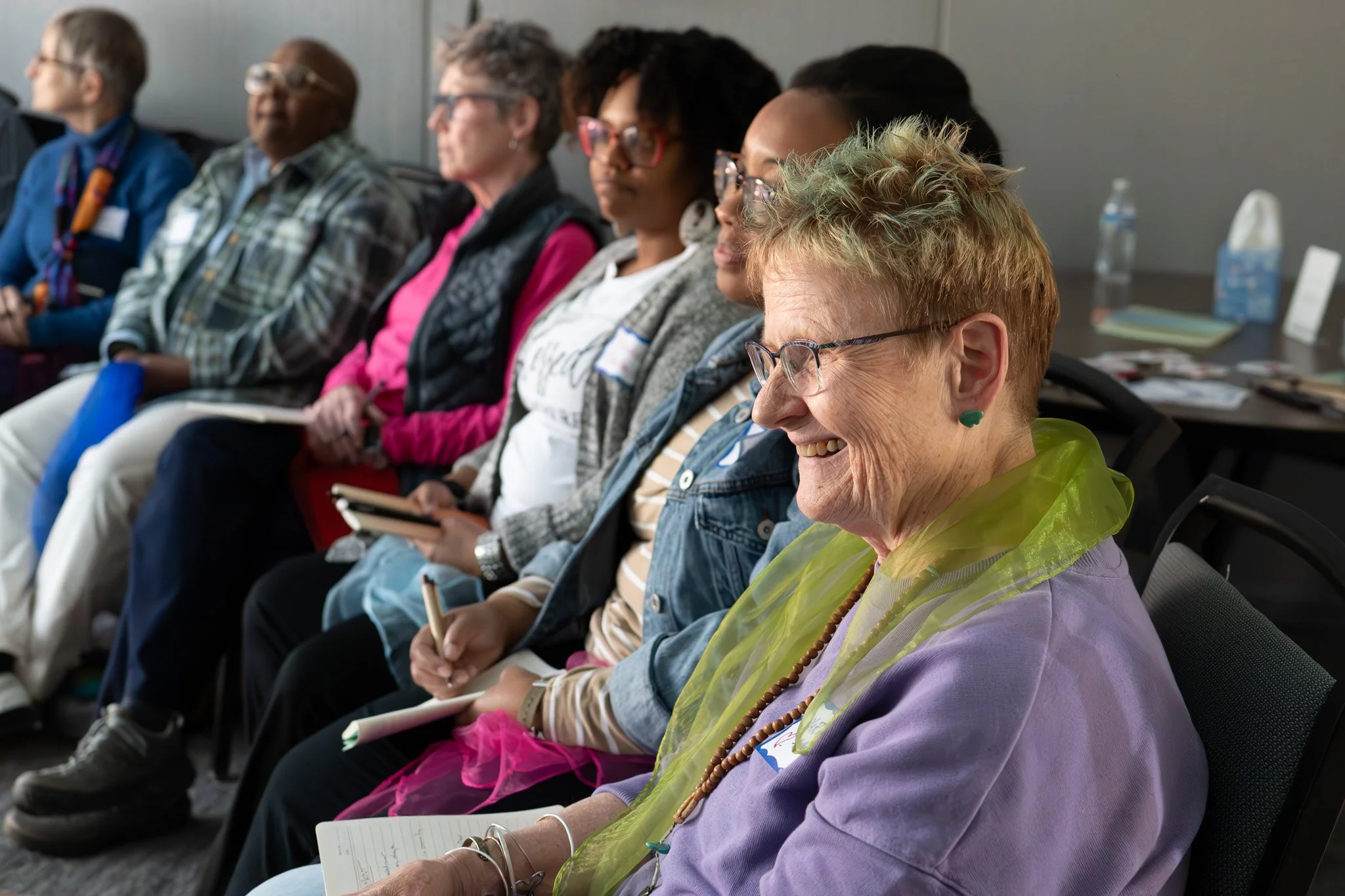A diverse group of people sitting in a conference room, listening attentively, with some taking notes and others smiling.