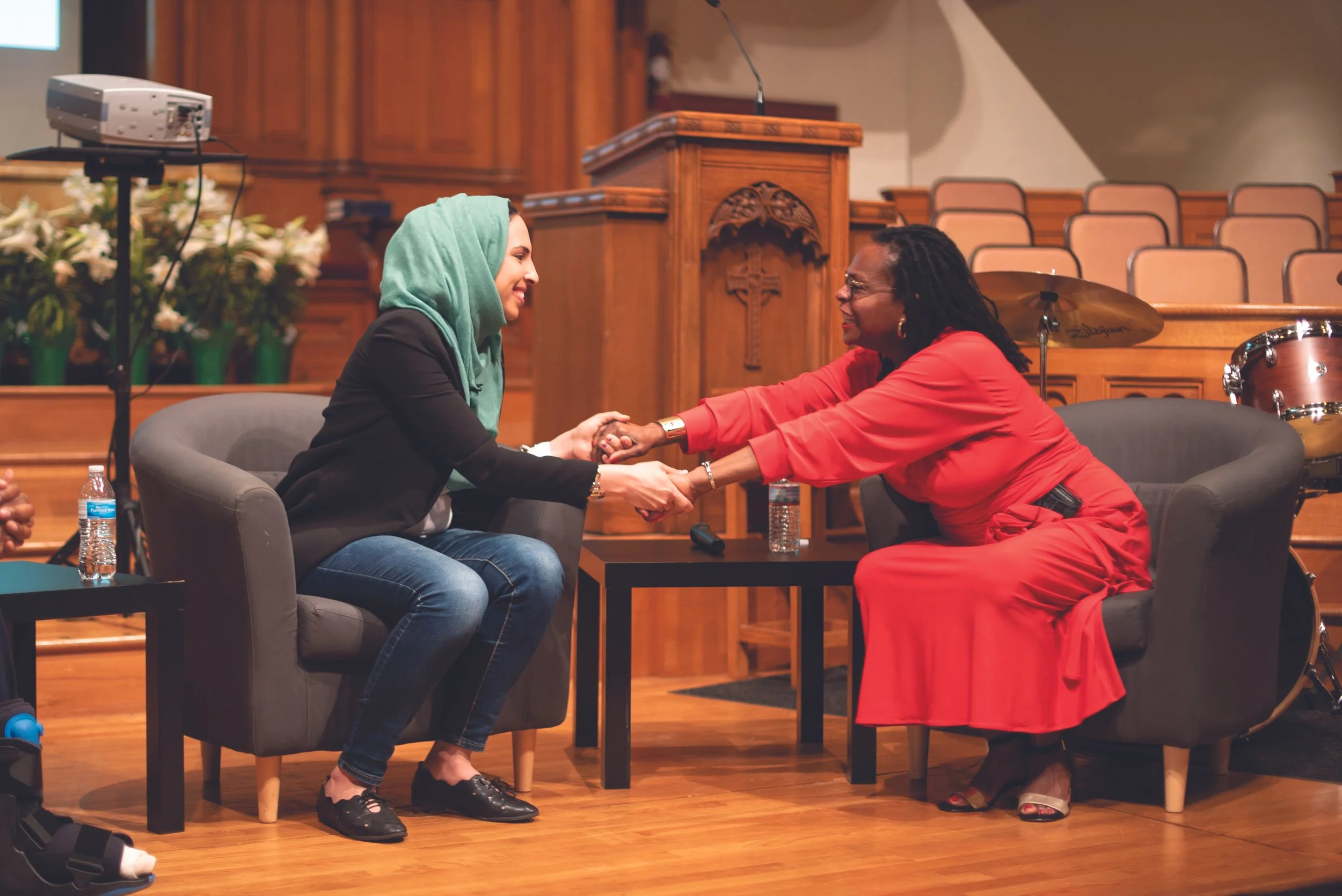 Two women sitting in chairs, smiling at each other and holding hands, in a church or auditorium setting with wooden accents and musical instruments in the background.