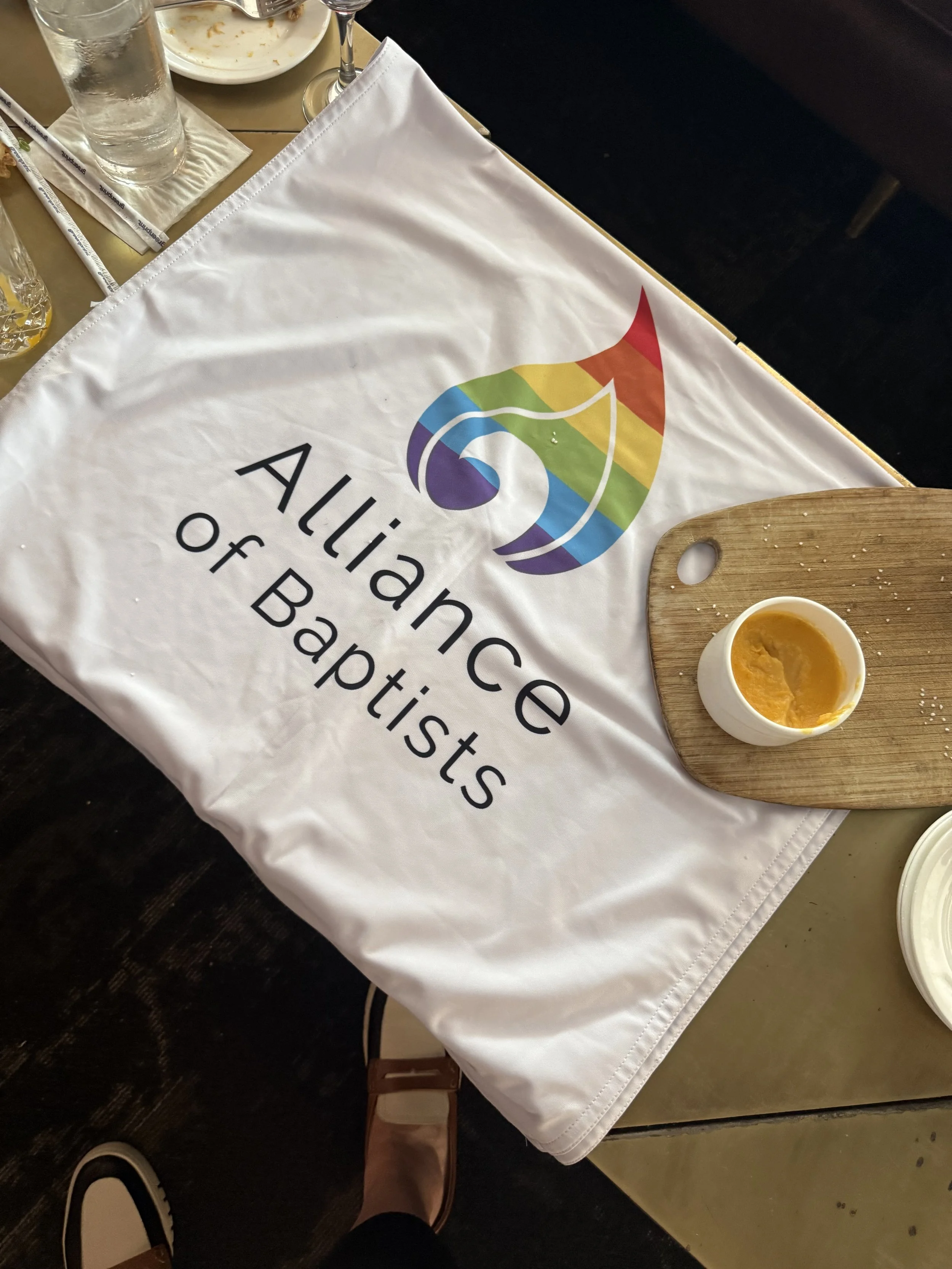 A table setting at a restaurant with a white cloth promoting the Alliance of Baptists. The table has a glass of water, a wine glass, a plate with leftover food, a cup of soup, chopsticks, and a small wooden cutting board with some crumbs.