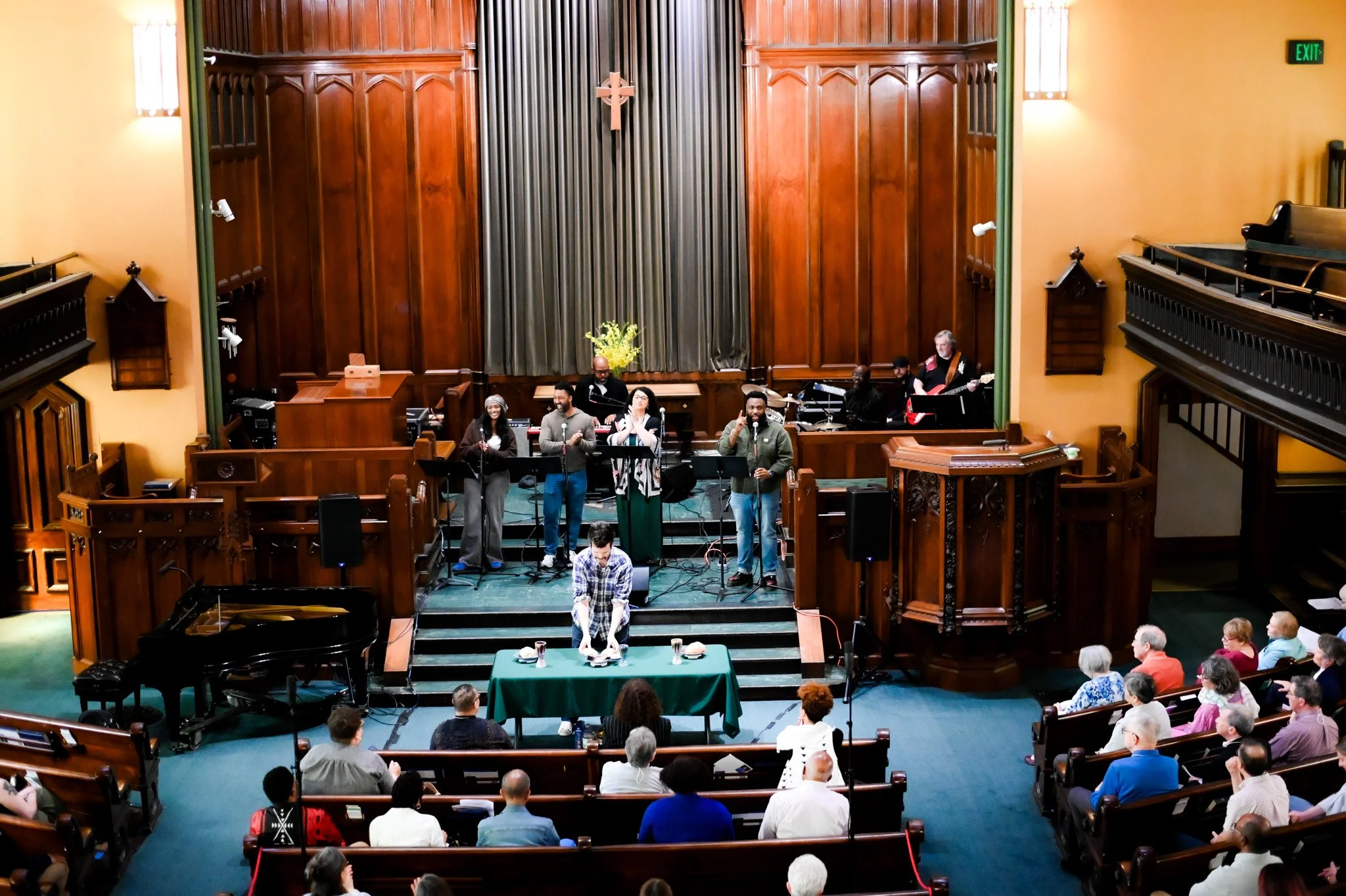 A group of people singing on a stage in a church sanctuary with wooden paneling, large curtains, and an audience seated in pews.