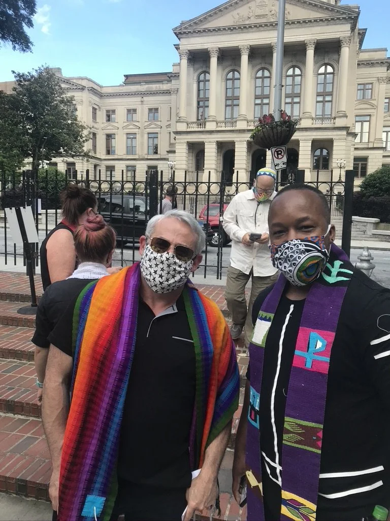 Two men wearing face masks and colorful scarves stand in front of a historic government building. Others are in the background, some also wearing masks, on a city street with bricks and parked cars.