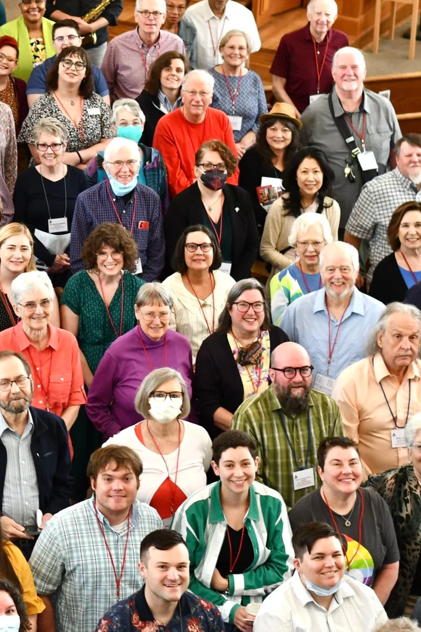 A large group of diverse people gathered indoors, many wearing name tags and smiling, posing for a group photo at an event or conference.