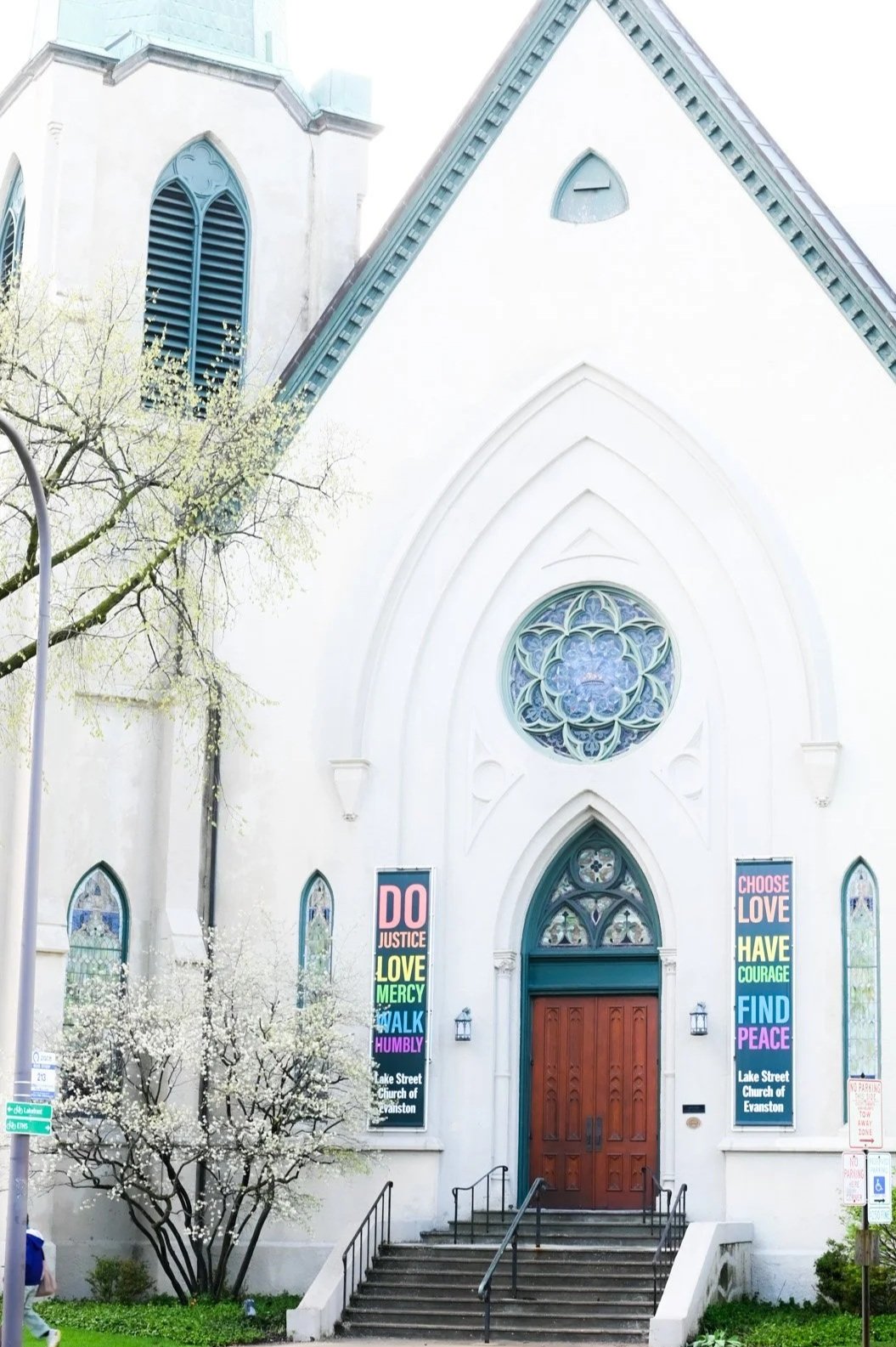 Front view of a white church with a pointed roof, stained glass windows, and colorful banners on either side of the entrance displaying words like 'Justice', 'Love', 'Peace', and 'Humble'. There are stairs leading up to a wooden door and blossoming trees nearby.