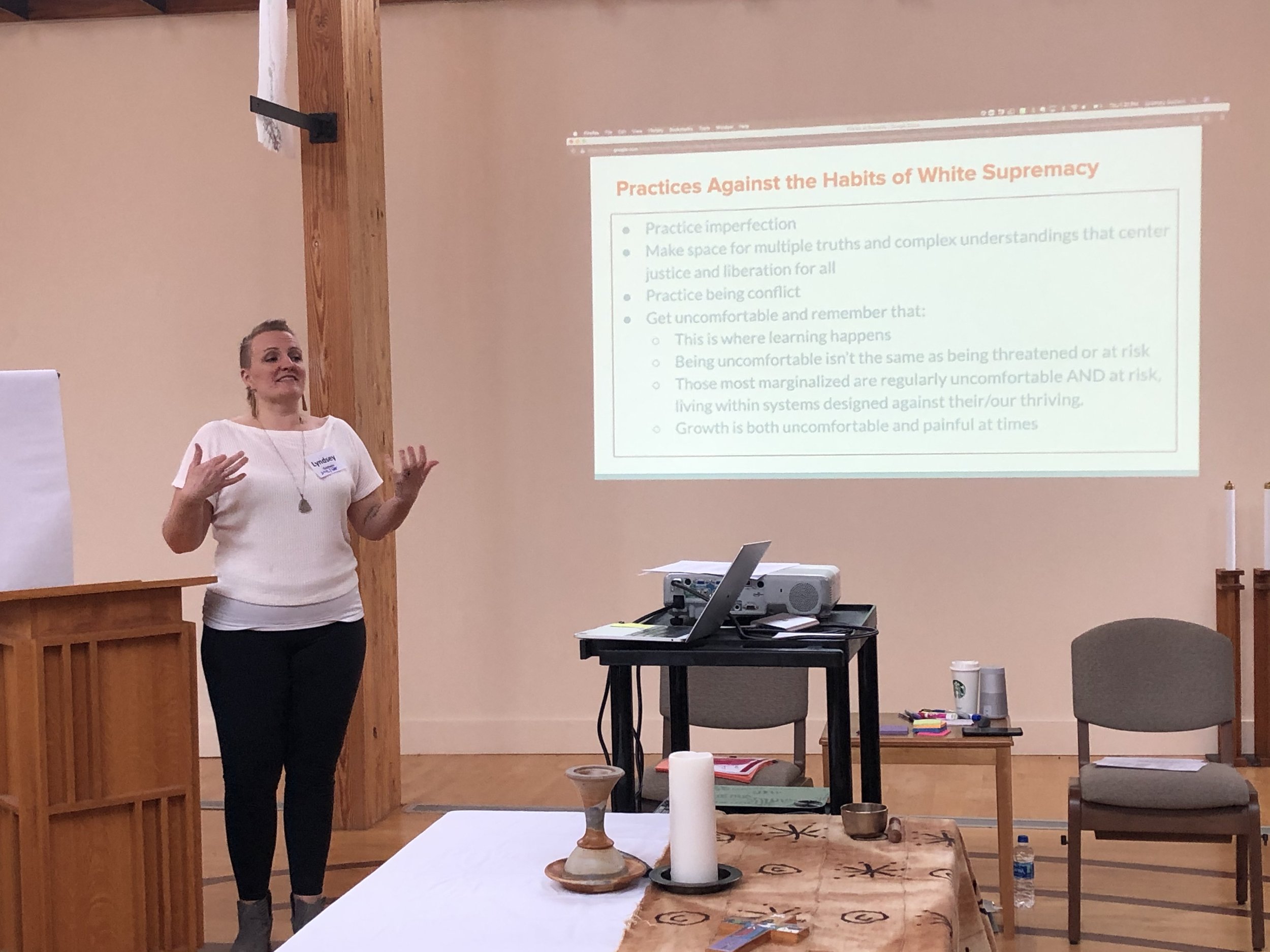 A woman giving a presentation in a room with a wooden wall and a large screen displaying a slide titled "Practices Against the Habits of White Supremacy." She is gesturing with her hands while speaking, and there are tables with various items, including candles, paper, and a water bottle, in front of her.