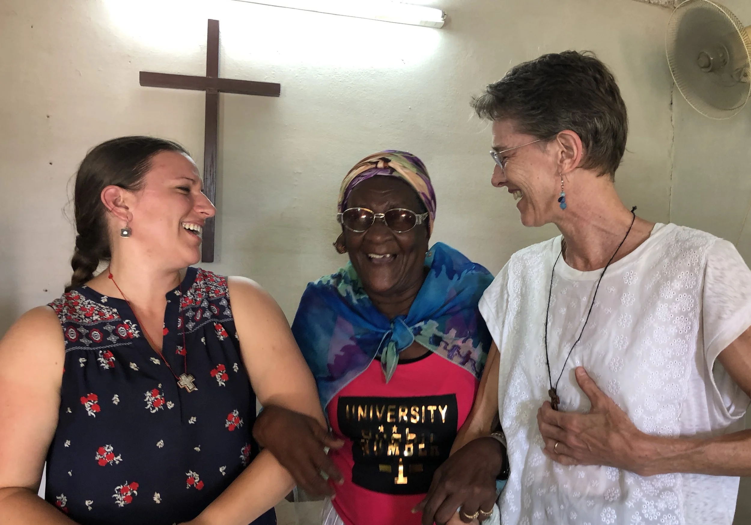 Three women laughing and smiling together indoors, with a cross on the wall and a ceiling fan in the background.