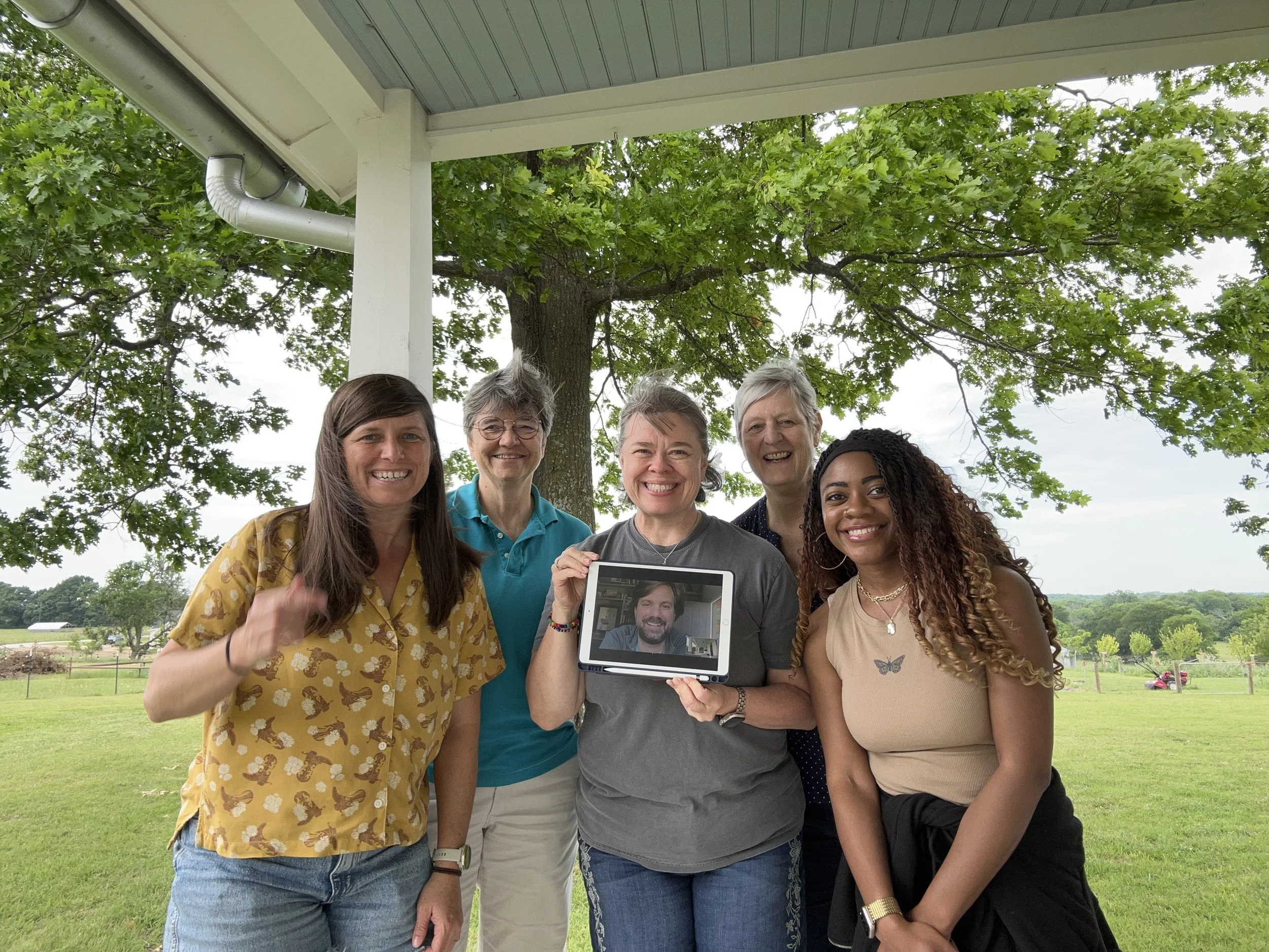 A group of six women standing outdoors in a park or garden, smiling at the camera, with green trees and grass in the background. One woman is holding a tablet showing a woman's face.