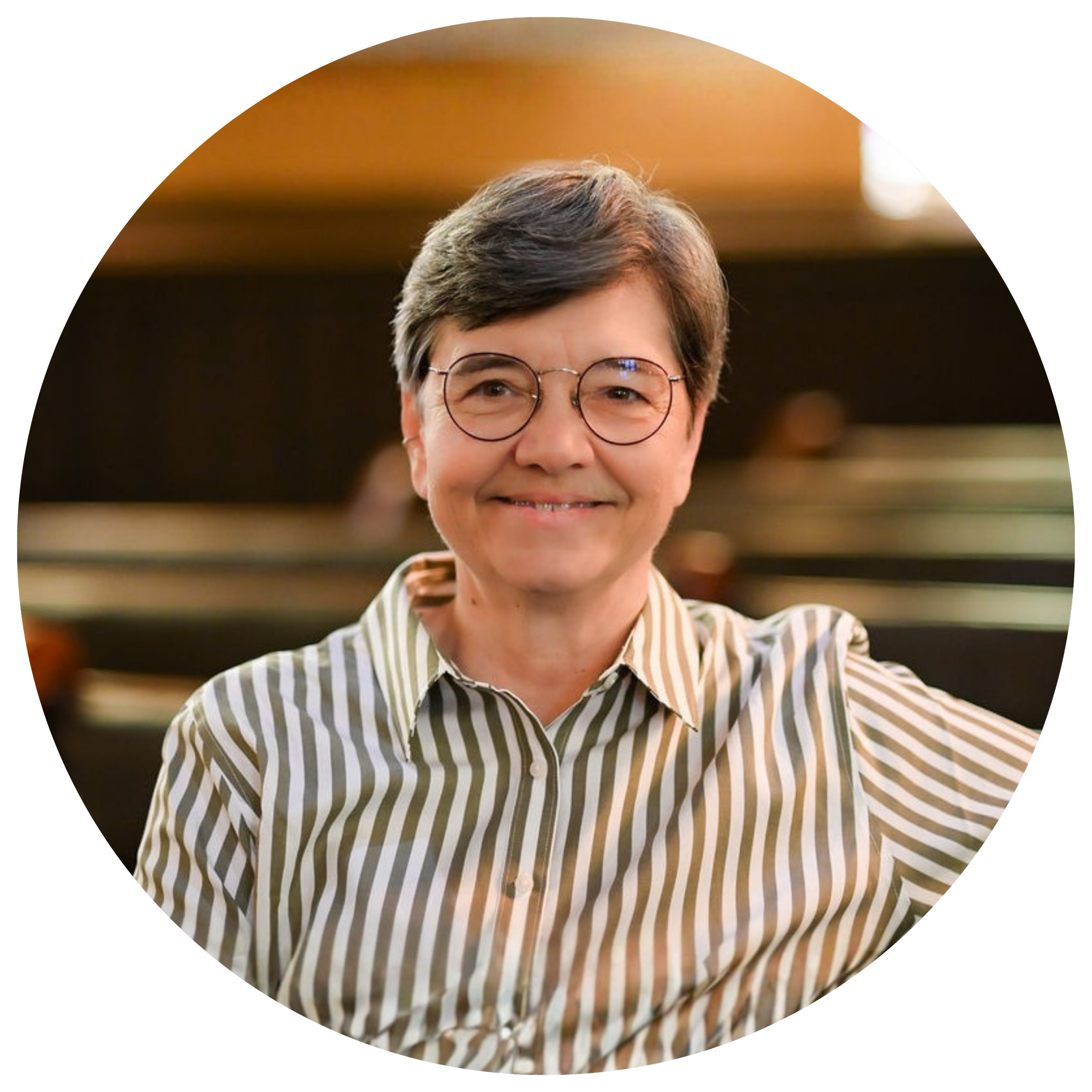 A woman with short gray hair and glasses smiling, wearing a striped shirt, in an indoor setting with warm lighting.