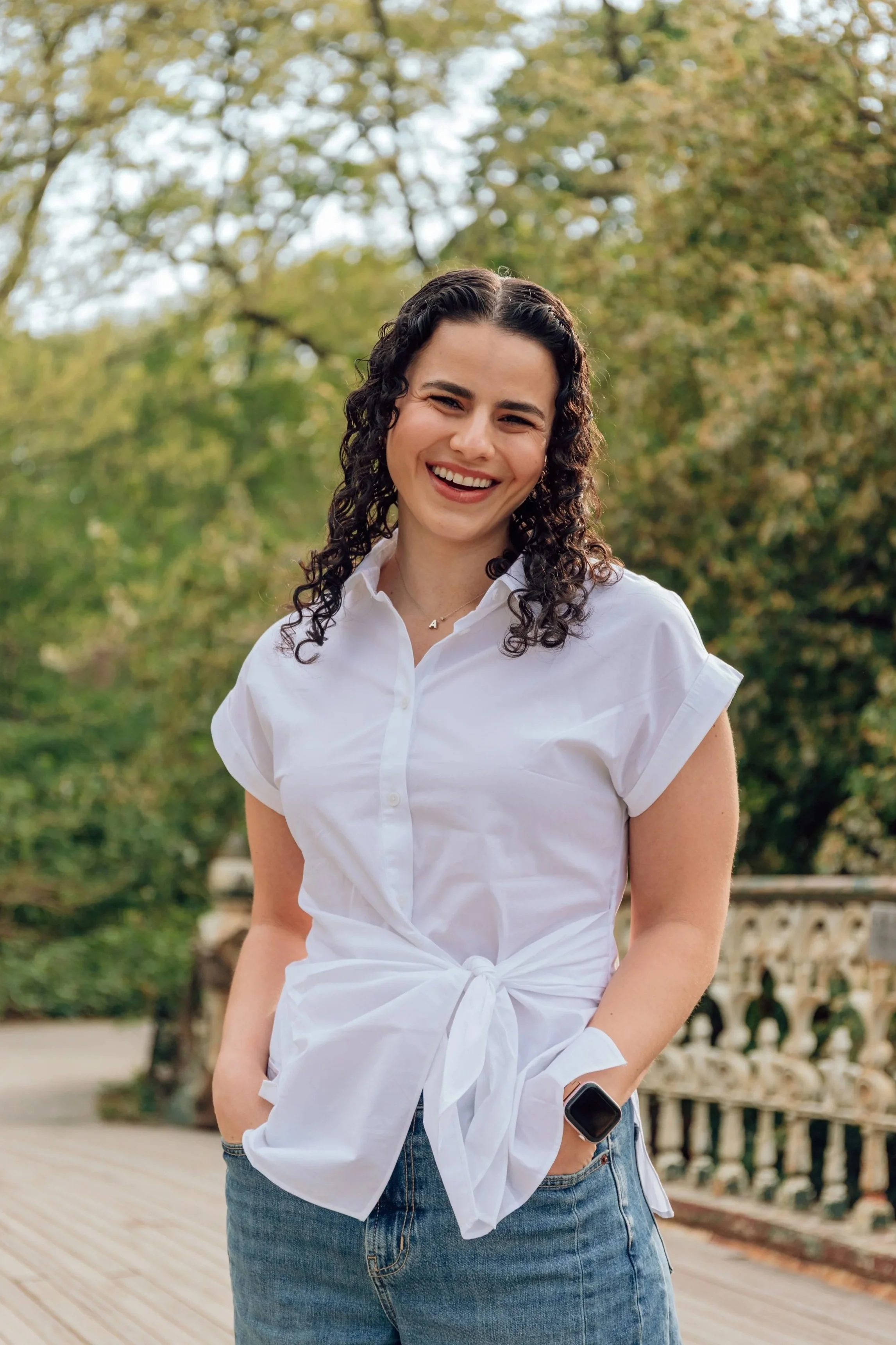 A young woman with curly dark hair smiling outdoors, wearing a white button-up shirt tied at the waist and jeans, standing on a wooden deck with trees in the background.