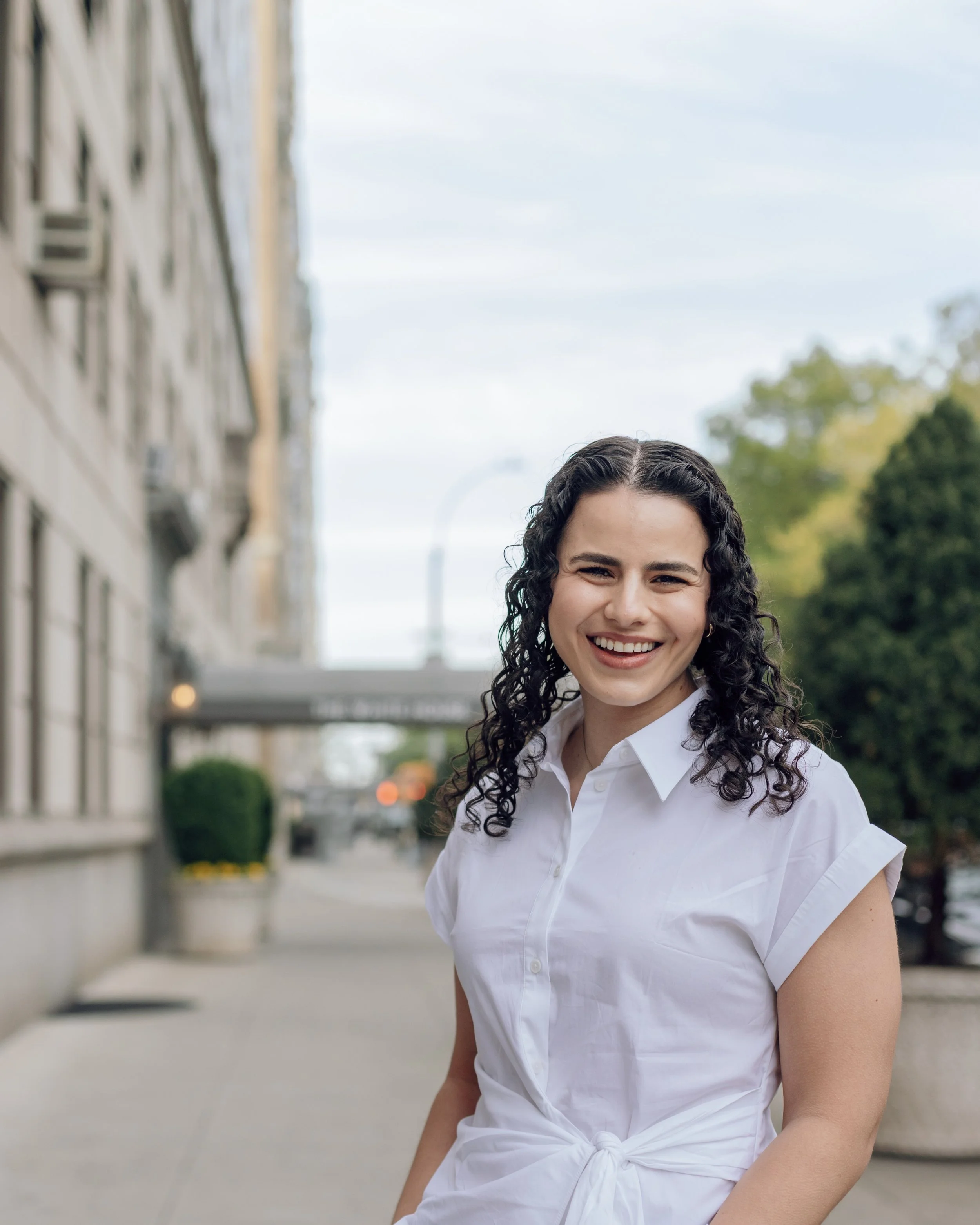 A woman with curly dark hair smiling outdoors in a white button-up shirt, standing on a city sidewalk with blurred buildings, trees, and a cloudy sky in the background.