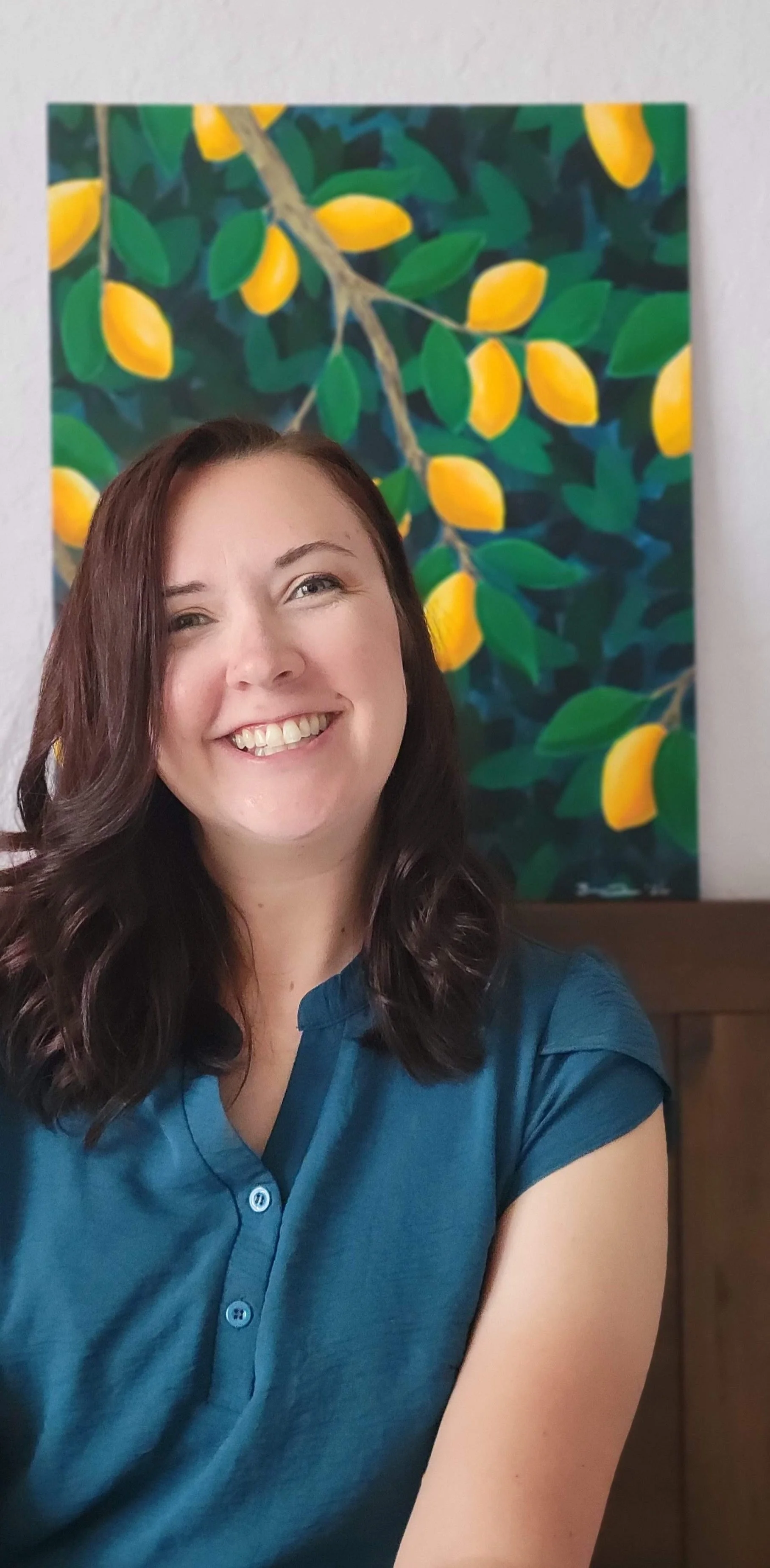 A woman with shoulder-length wavy brown hair and a blue shirt smiling in front of a lemon-themed painting on a white wall.