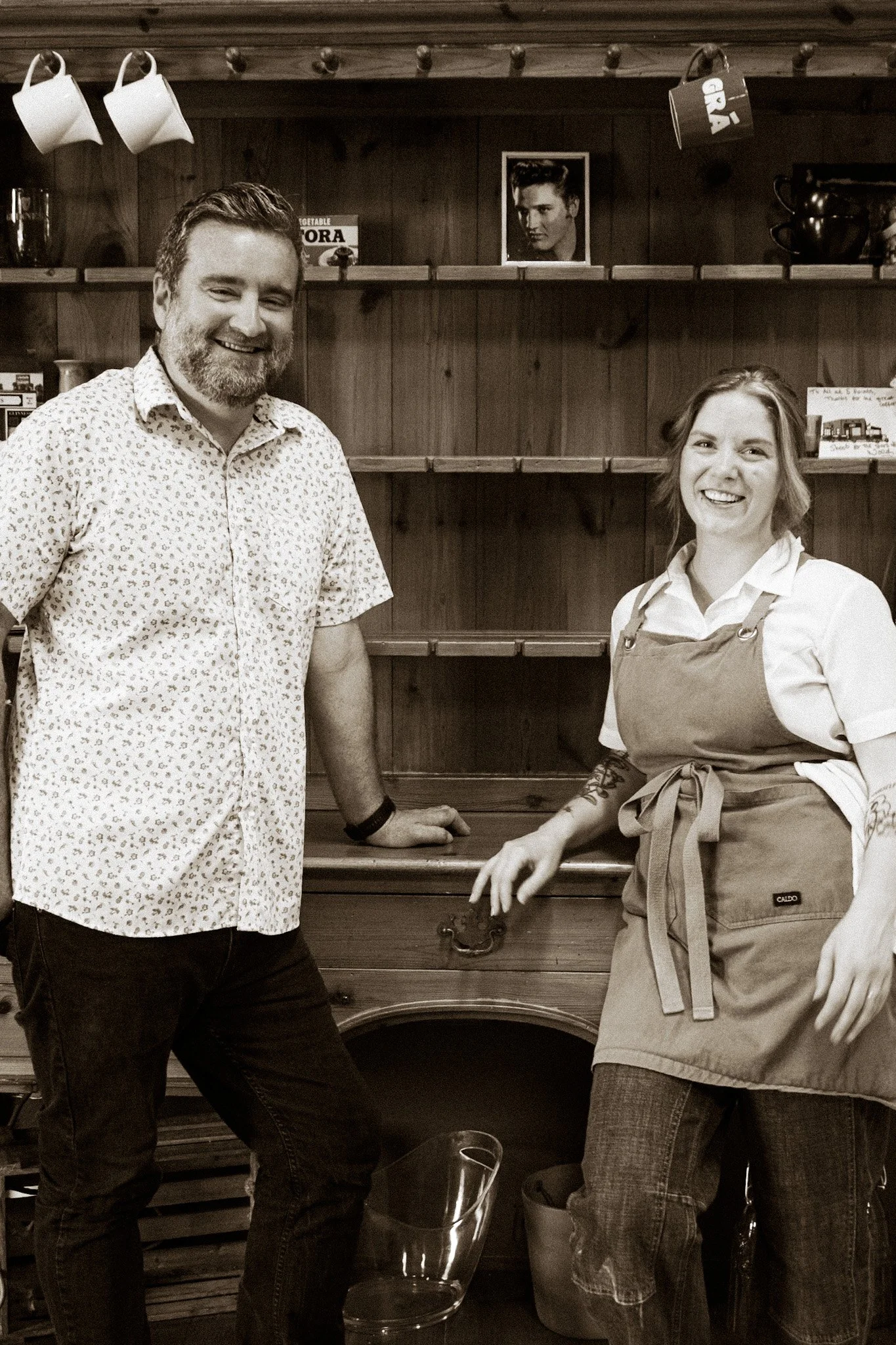 Two people standing in front of a wooden background with shelves. The man on the left has a beard, is smiling, and wearing a patterned short-sleeved shirt. The woman on the right has short hair, is smiling, and wearing a white shirt with an apron. Th