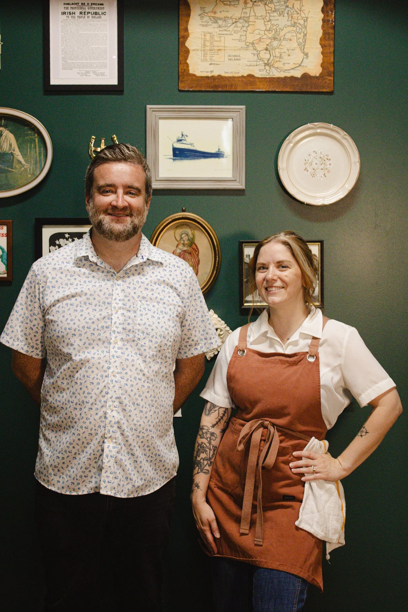 A man with a beard wearing a white shirt with a floral pattern and a woman with a tattooed arm wearing a brown apron over a white shirt, standing in front of a wall decorated with framed pictures, including a ship, religious figures, and maps.