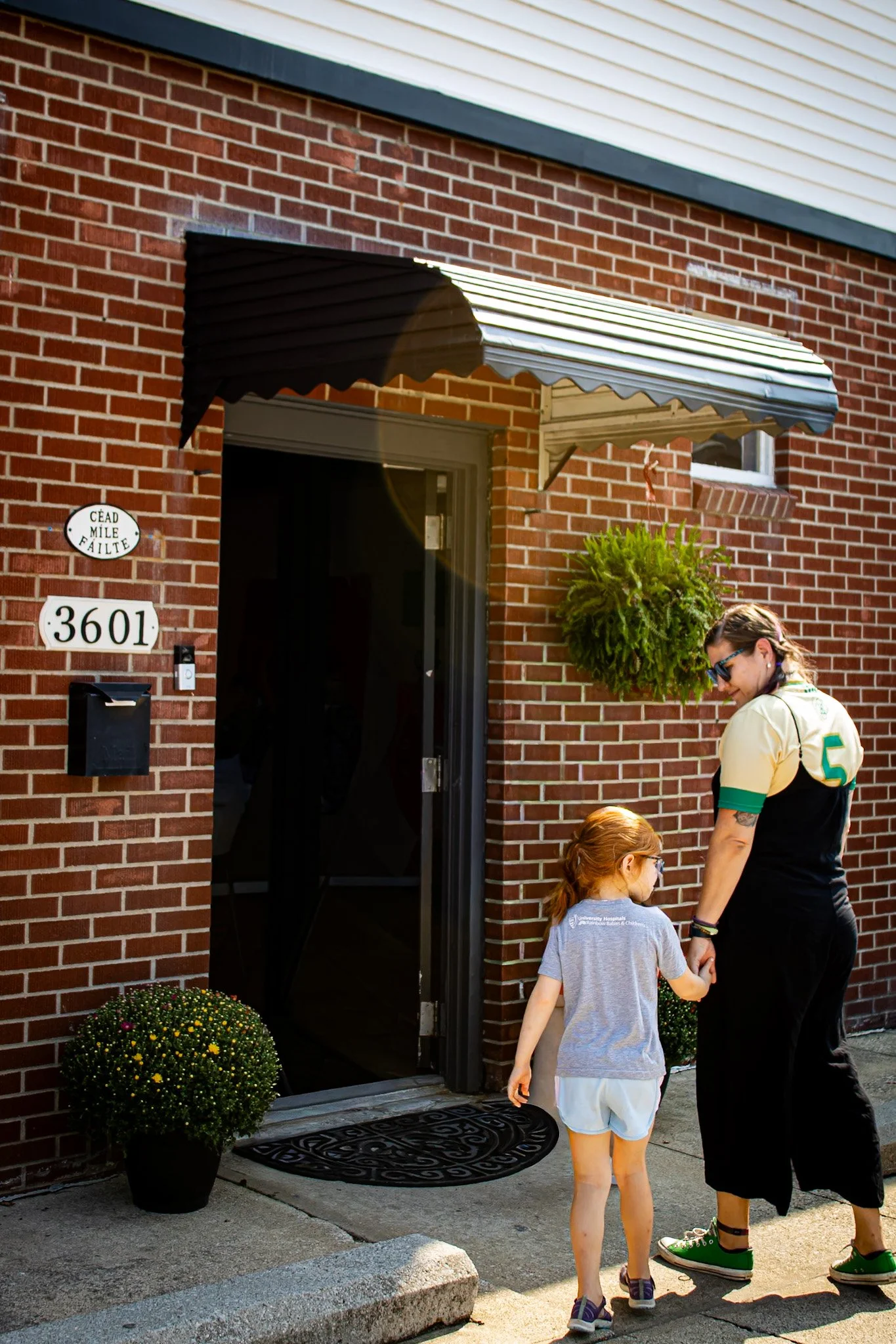 A woman and a young girl holding hands outside a brick house with a black door and awning. There are yellow and white flowers in pots on each side of the steps. The woman is wearing a sports jersey, sunglasses, and green shoes. The girl has red hair tied back and is dressed in a grey shirt and shorts.