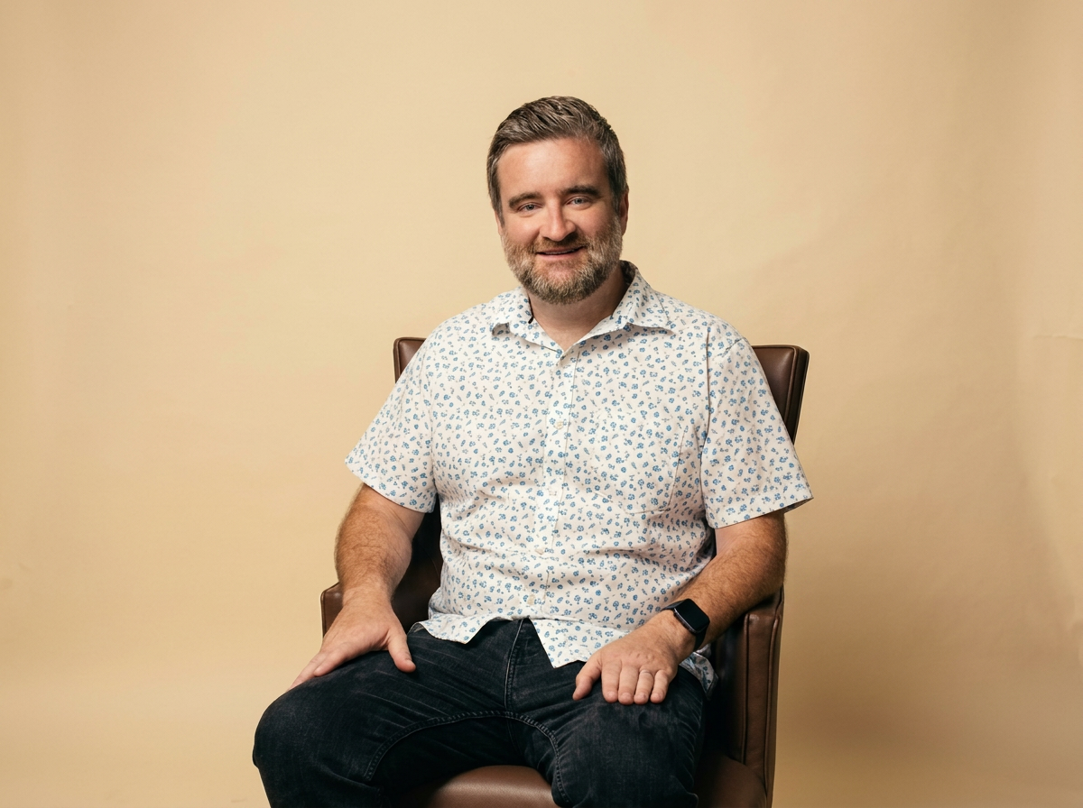 A smiling man with a beard and short brown hair, wearing a white short-sleeve shirt with blue floral patterns, black jeans, and a smartwatch, sitting on a brown leather chair against a plain beige background.