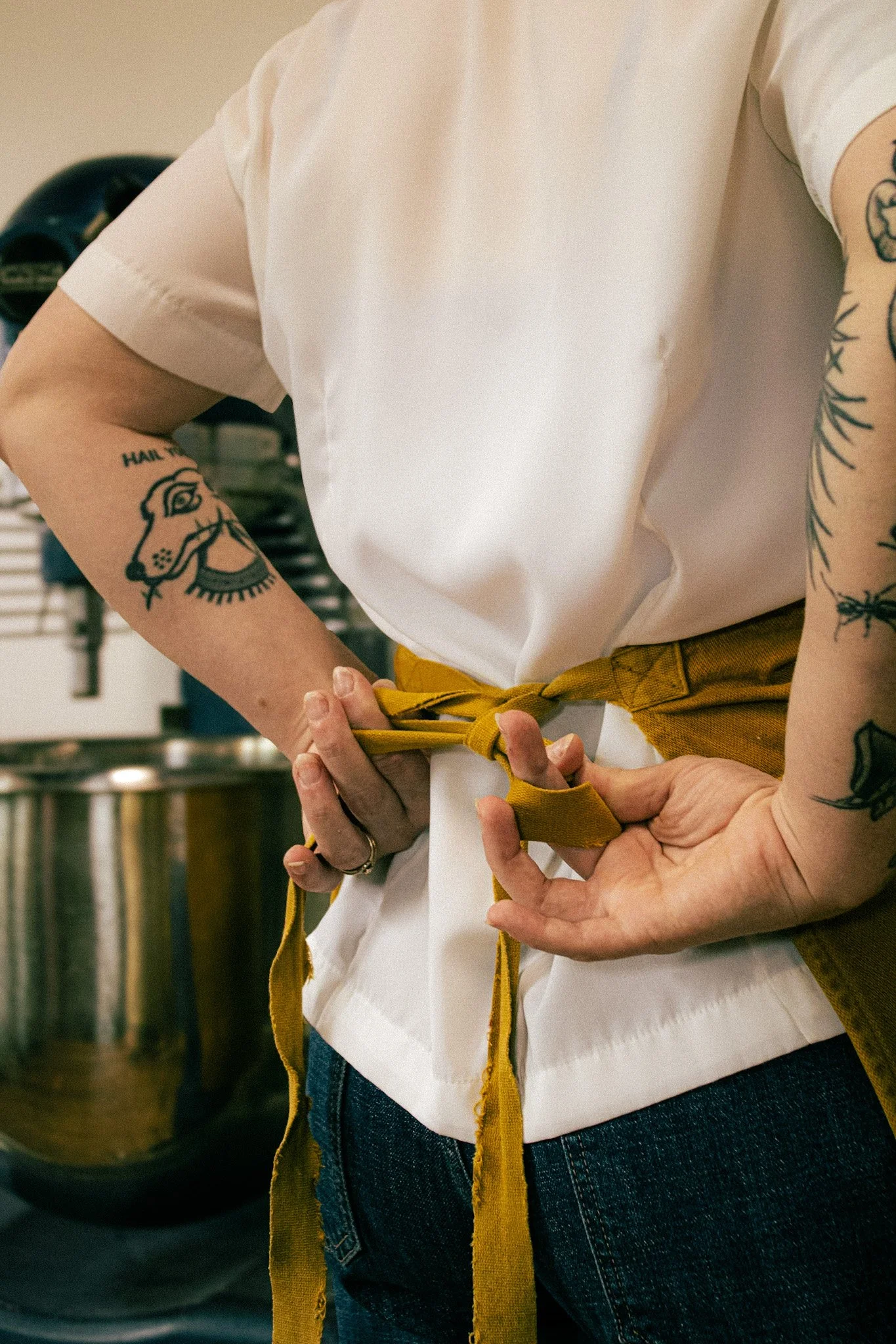 Close-up of two people tying yellow apron strings, one person is wearing a white shirt with tattoos on both arms, one tattoo is a face of a lion, and there is a washing machine in the background.