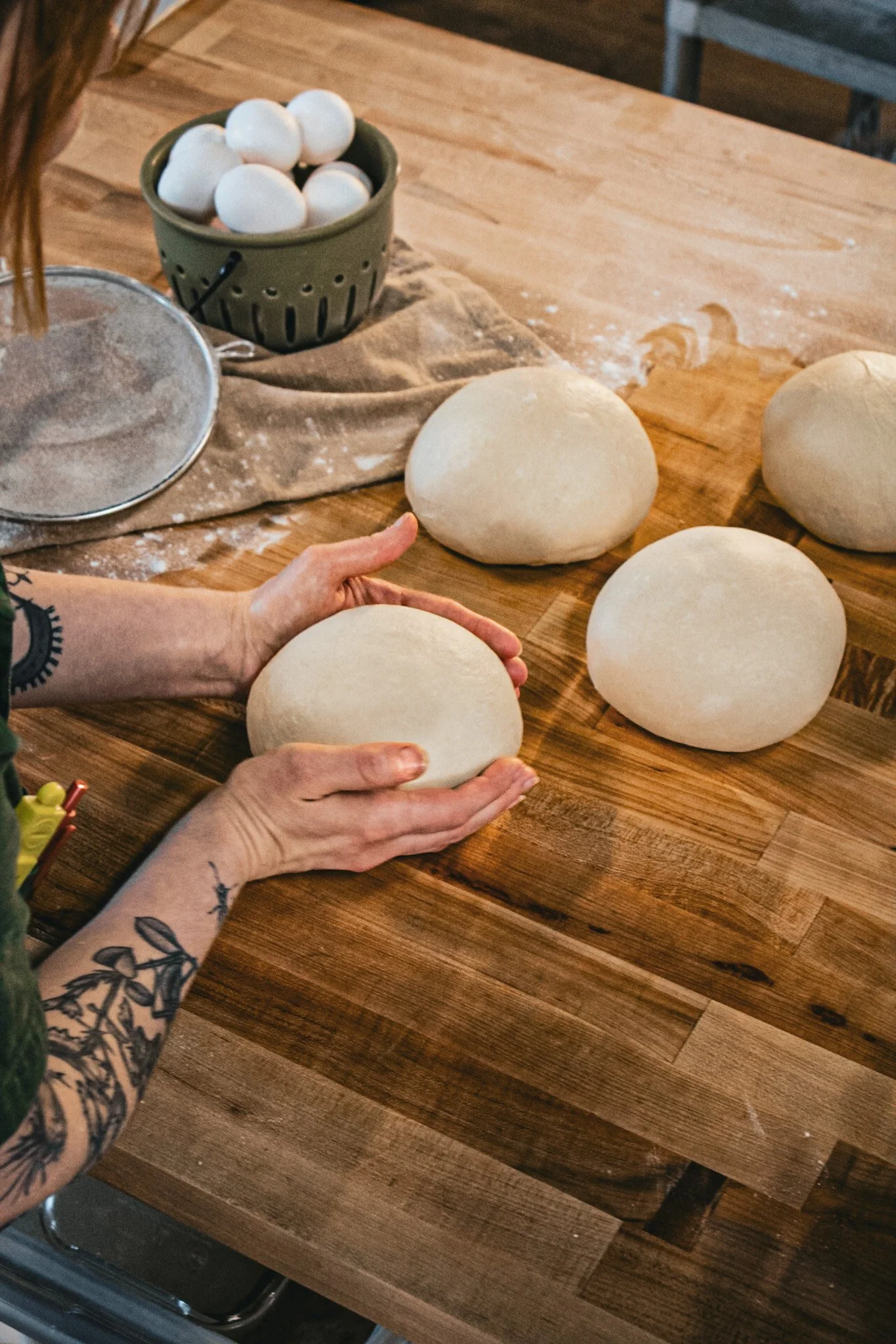 Person with tattooed arms handling a ball of dough on a wooden table with more dough balls, a sieve, and a bowl of eggs in the background.