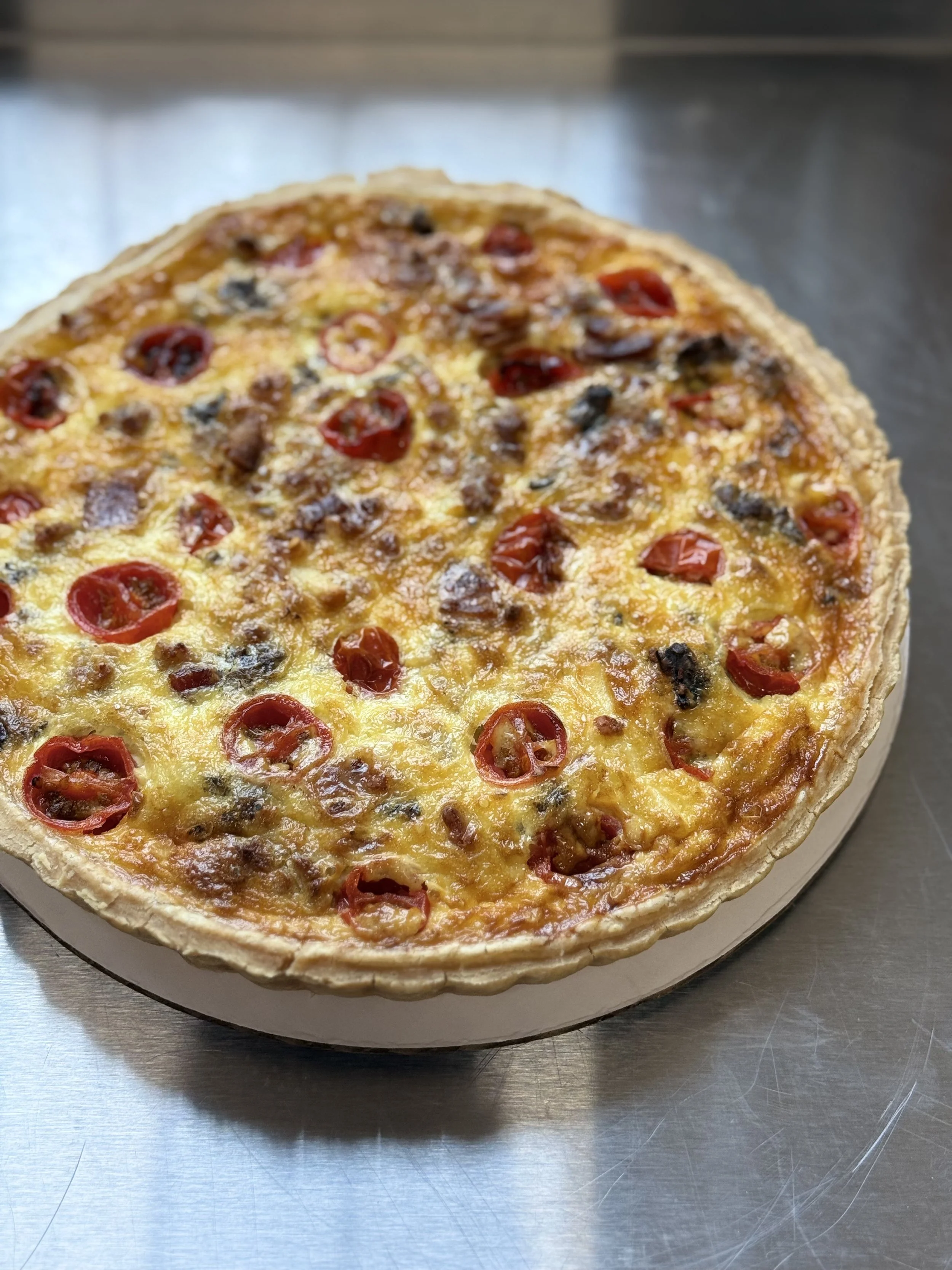 Whole deep dish pizza topped with cherry tomatoes, sausage, and black olives, on a stainless steel surface.