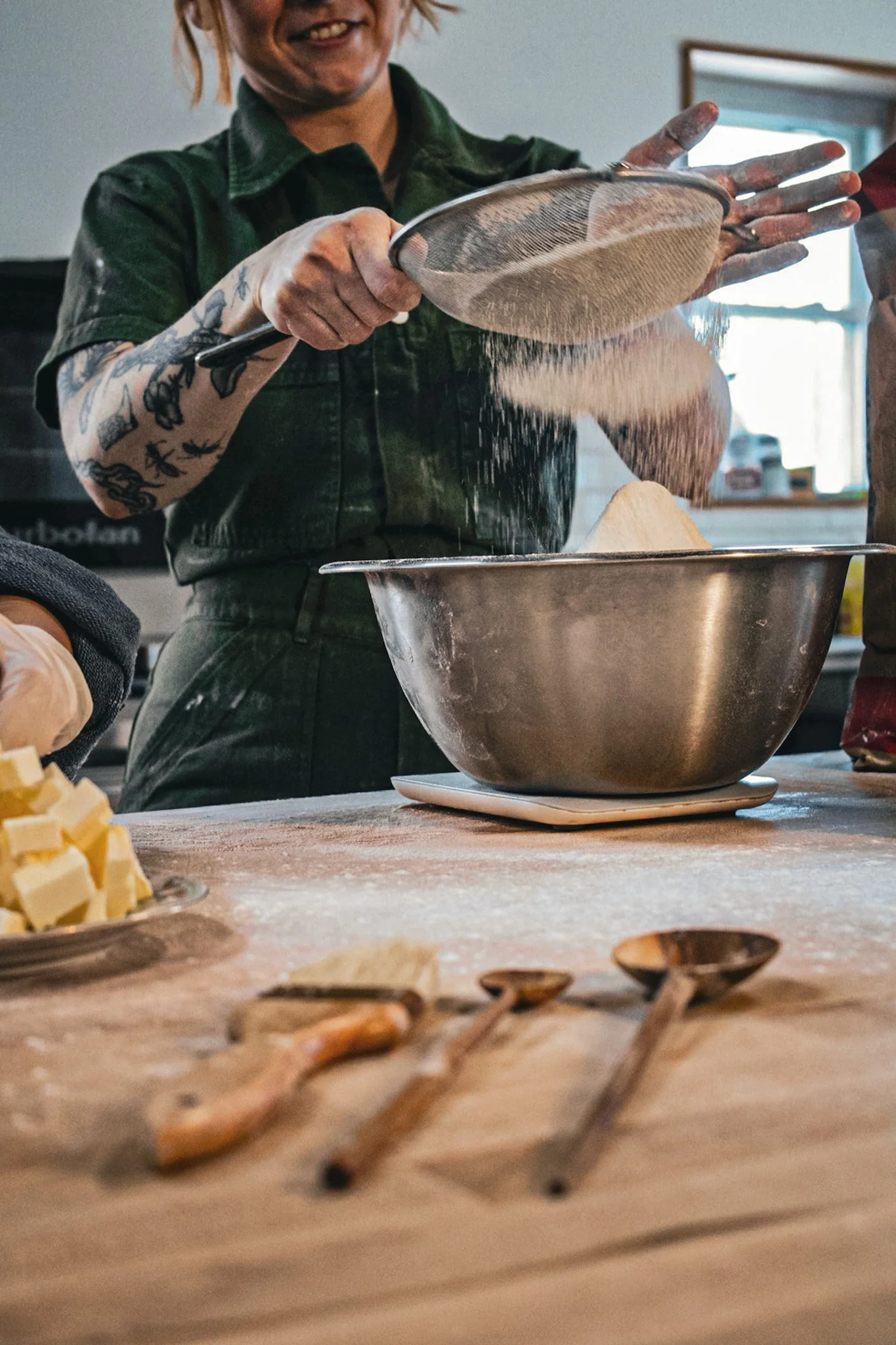 A person with tattoos on their arm sifting flour into a mixing bowl in a kitchen. The person's face is partially visible, showing a smile. There are ingredients and utensils on a wooden countertop with some baking tools and ingredients partially visi