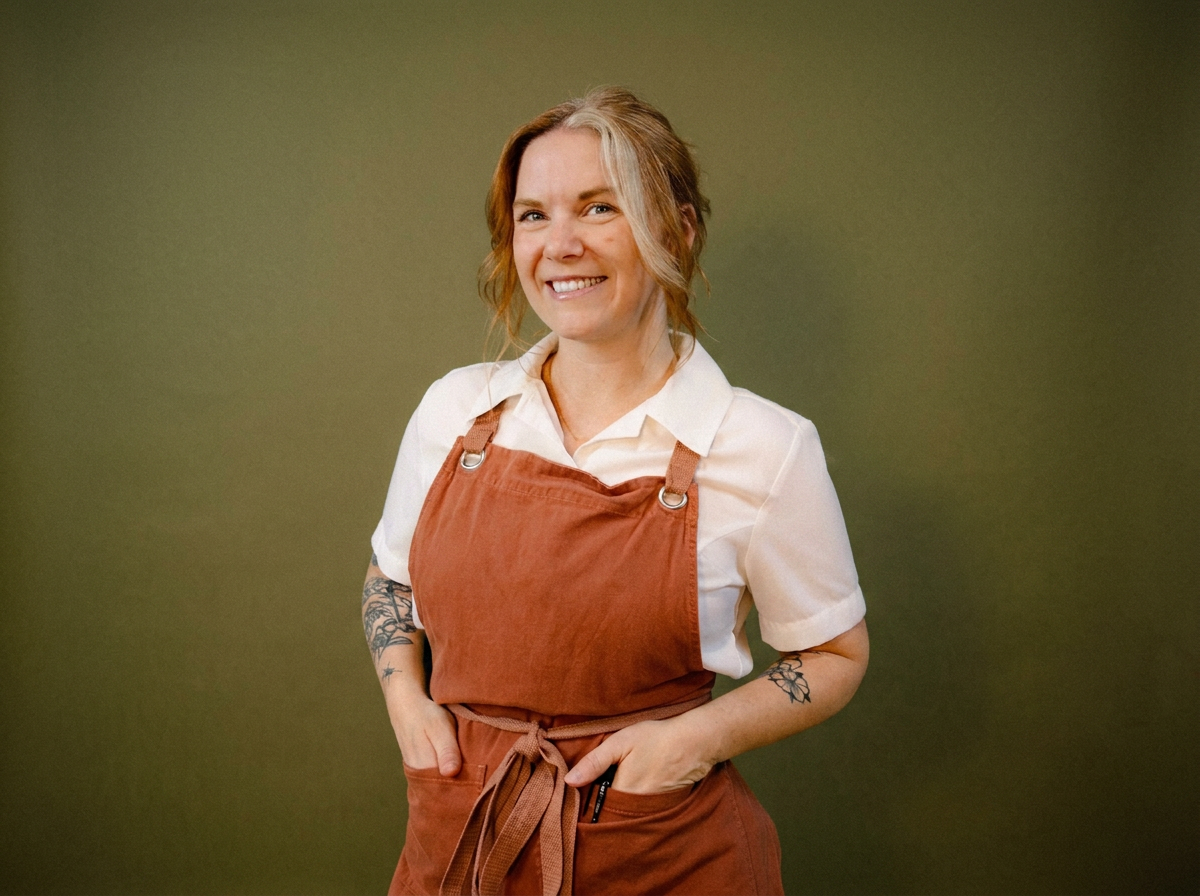 A smiling woman with tattoos on her arms, wearing a white shirt and an orange apron, standing against a green background.